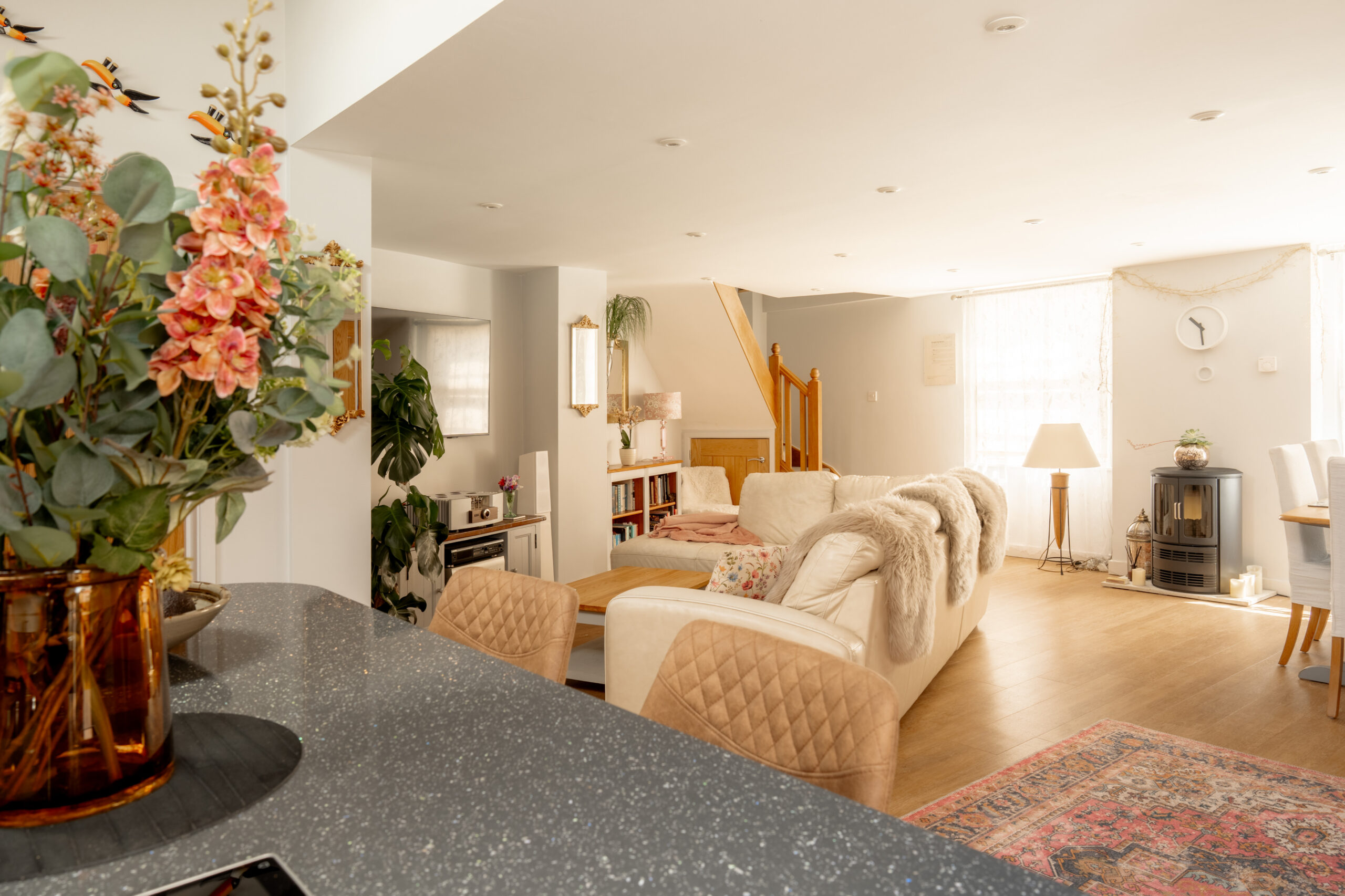 Bright, cozy living room with a white sofa, armchairs, and wooden floors. Large windows let in natural light. A vase with flowers sits on a counter in the foreground. Stairs and shelves are visible in the background.