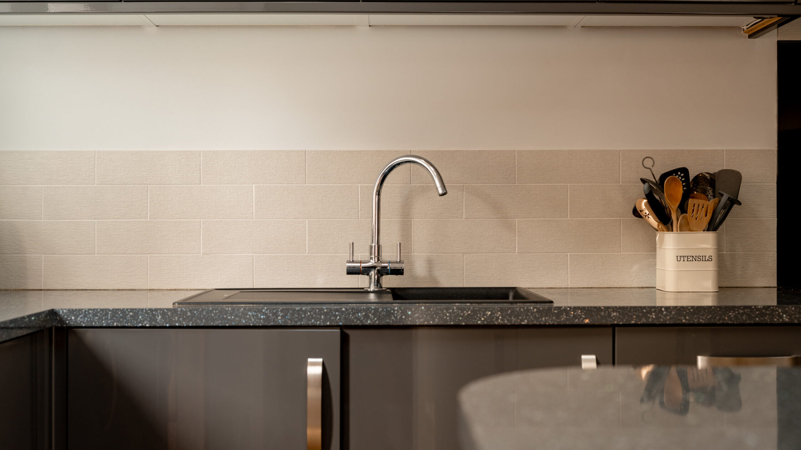 Modern kitchen with glossy dark cabinets, chrome handles, a stainless steel faucet, light tile backsplash, and a white container holding utensils on the right side of the countertop.