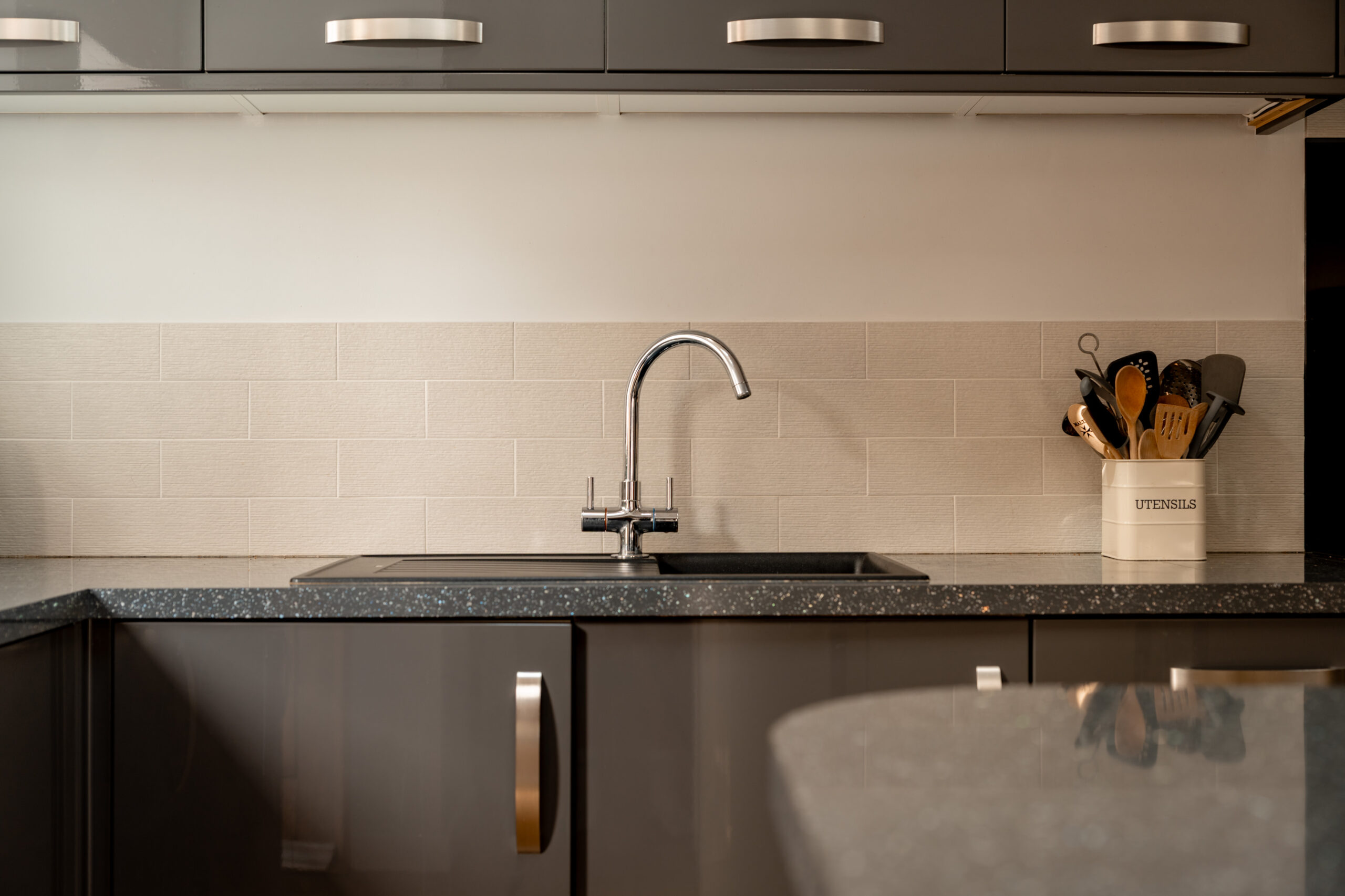 Modern kitchen with glossy dark cabinets, chrome handles, a stainless steel faucet, light tile backsplash, and a white container holding utensils on the right side of the countertop.