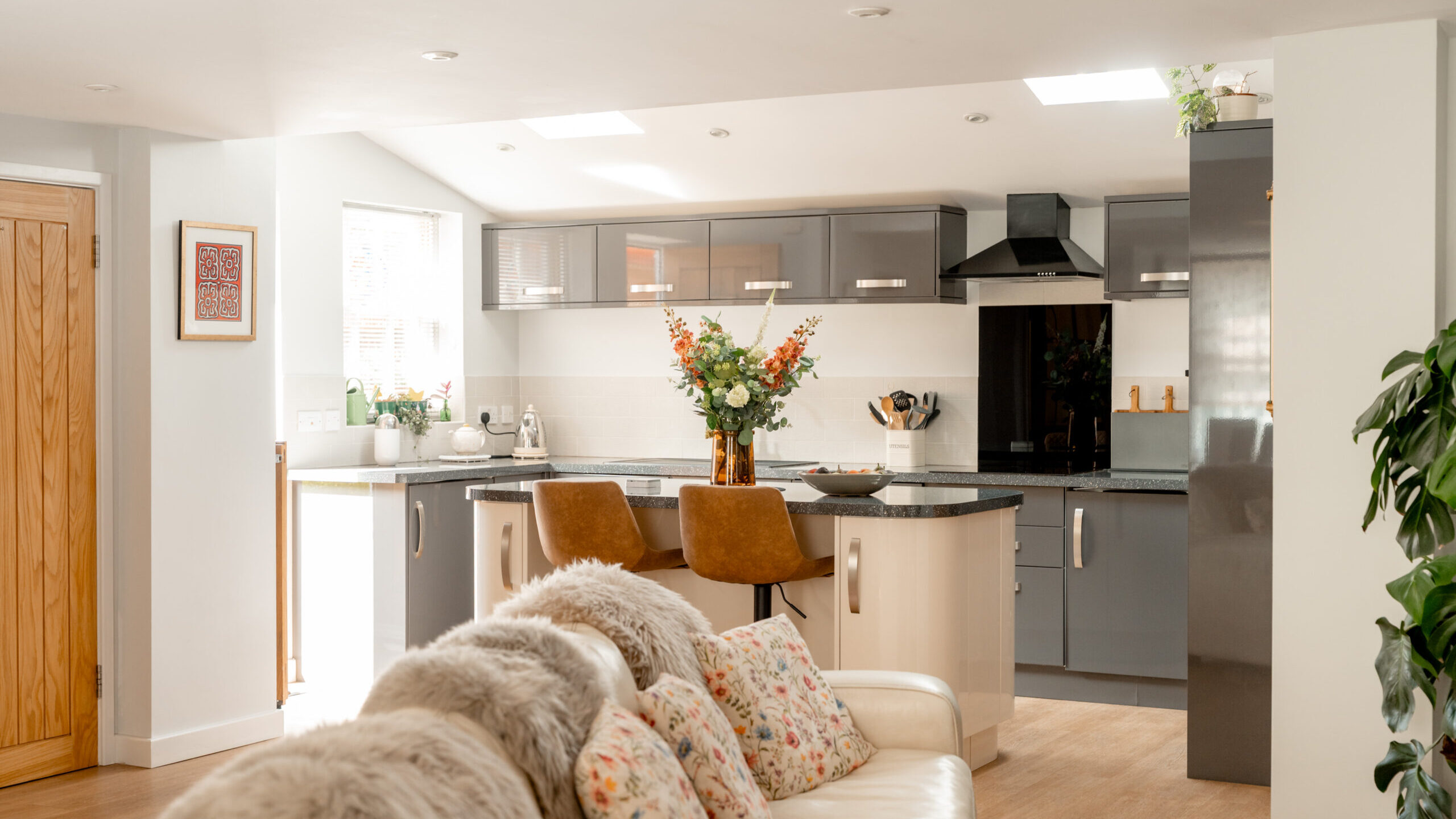 Modern kitchen with gray cabinets, an island with two brown chairs, fresh flowers, and a cozy living area featuring a white sofa with floral cushions and fur throws. Bright natural light fills the space.