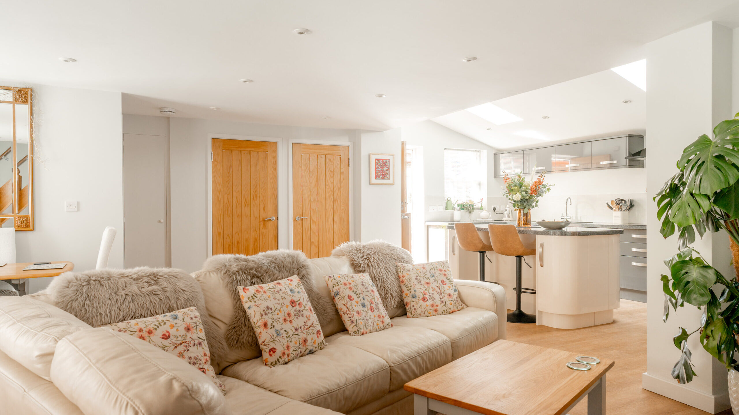 Bright, modern open-plan living room and kitchen with beige sofa, floral cushions, wooden coffee table, breakfast bar with stools, and a large green plant. Natural light fills the space through skylights.