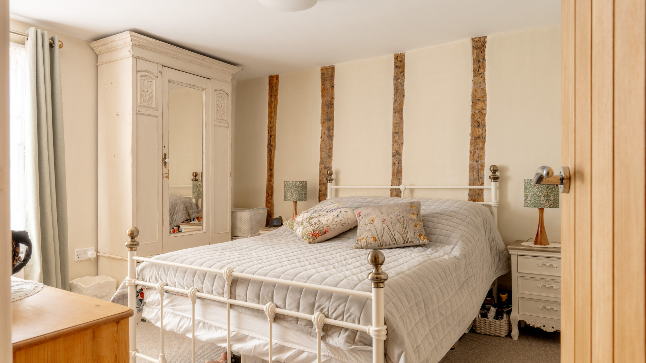 Cozy bedroom with a white metal bed, patterned pillows, vintage wardrobe with mirrors, bedside tables with lamps, and exposed wood accents on the wall. Sunlight streams in through a window with curtains.