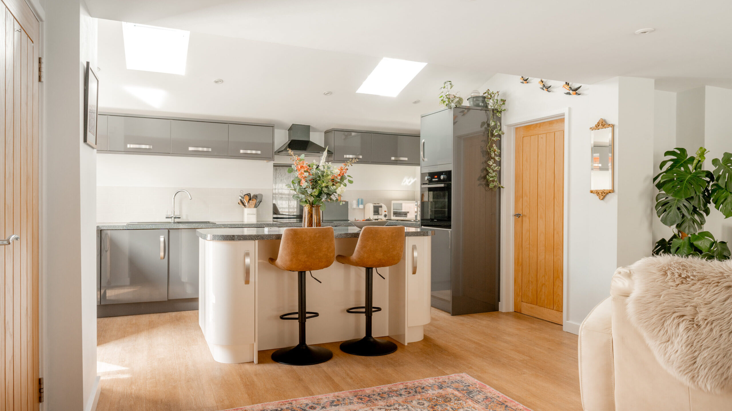 Modern kitchen with gray cabinets, a white island with two tan barstools, plants, and a colorful rug. Skylights and wood accents create a bright, cozy atmosphere.