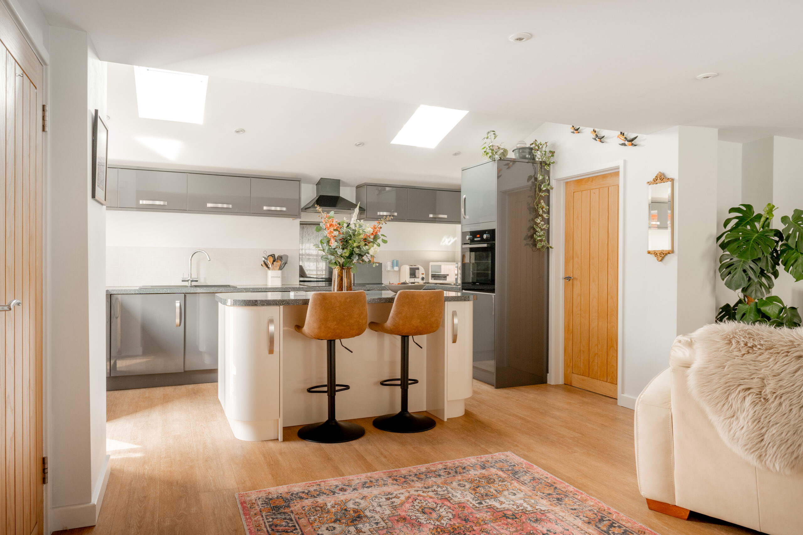 Modern kitchen with gray cabinets, a white island with two tan barstools, plants, and a colorful rug. Skylights and wood accents create a bright, cozy atmosphere.