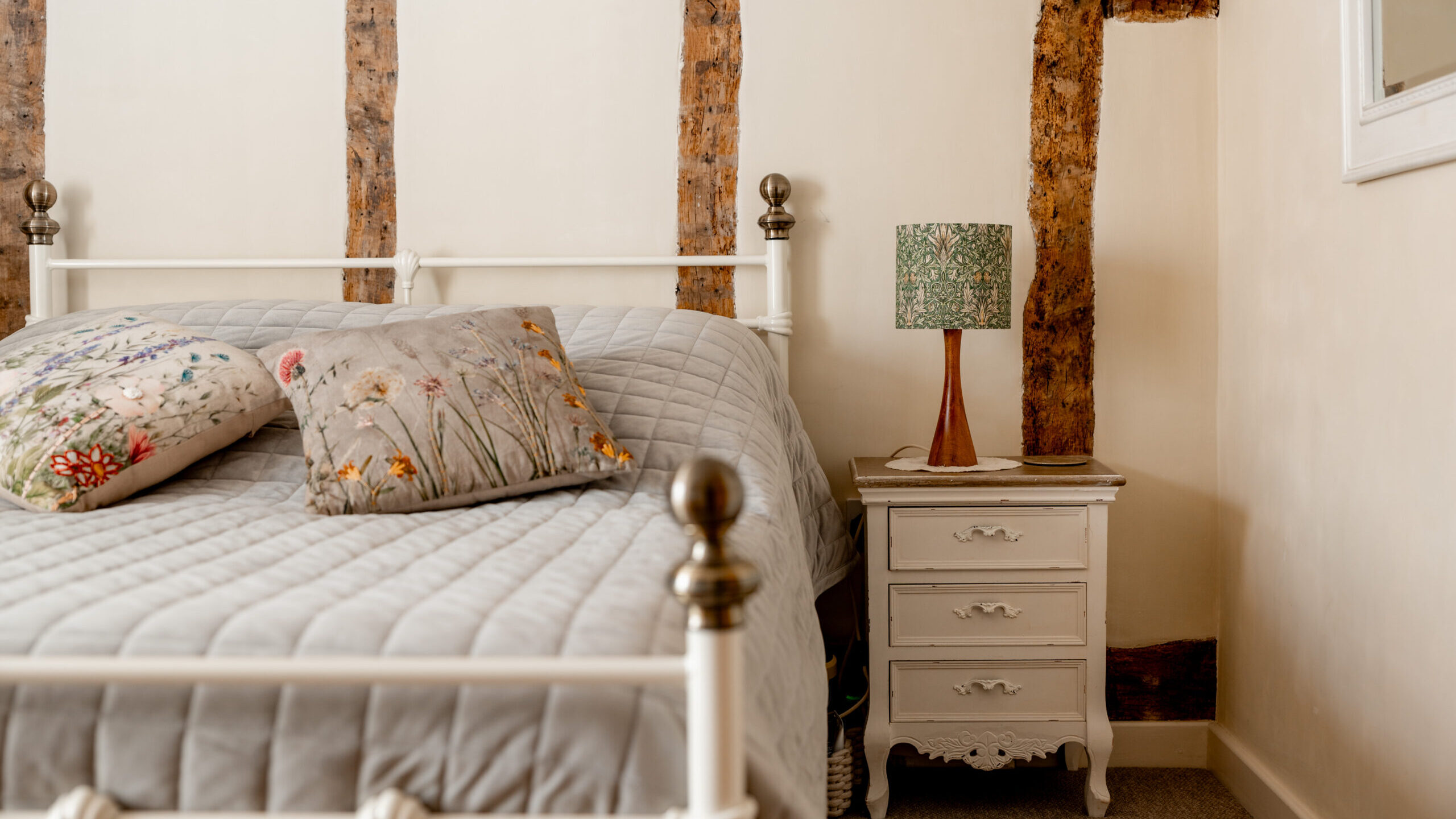 A cozy bedroom with a white metal bed, floral pillows, a gray quilt, a small white nightstand, and a patterned lamp. Wooden beams are visible on the cream-colored wall.