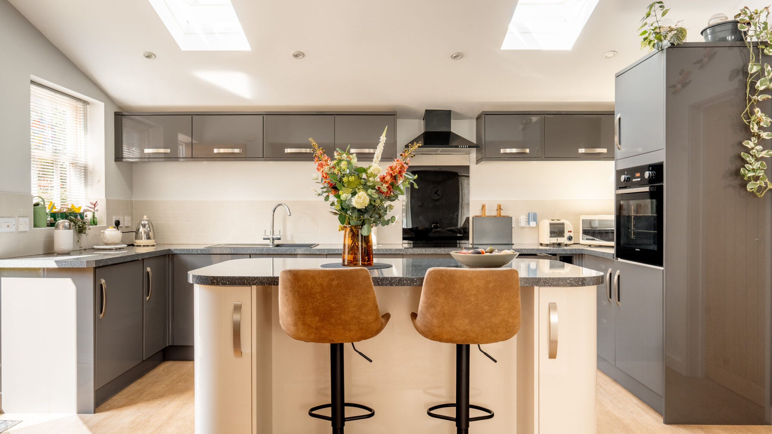 Modern kitchen with grey cabinets, white countertops, two brown barstools at an island, a vase of flowers, and natural light from skylights and a window. Appliances and plants decorate the space.