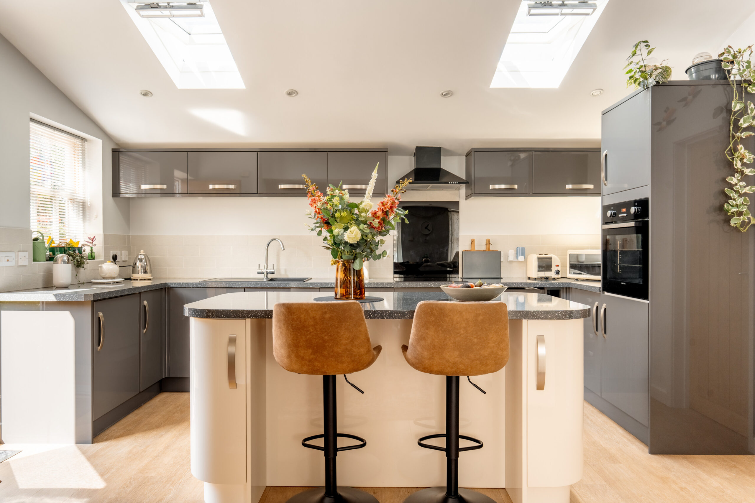 Modern kitchen with grey cabinets, white countertops, two brown barstools at an island, a vase of flowers, and natural light from skylights and a window. Appliances and plants decorate the space.