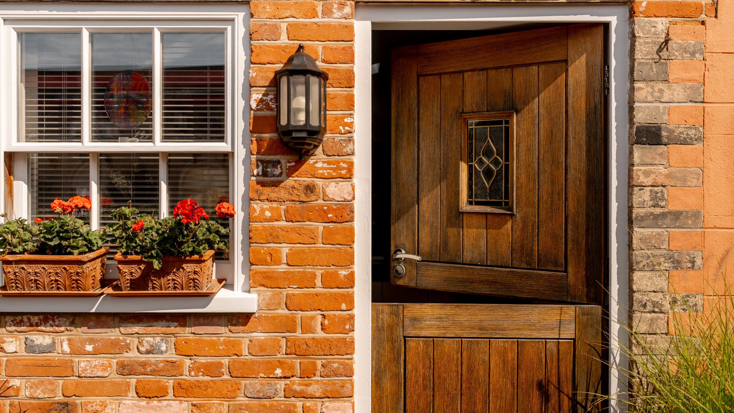 A partially open wooden door on a brick house, next to a window with white trim and flower boxes filled with red and orange flowers. A black wall lantern is mounted between the door and window.