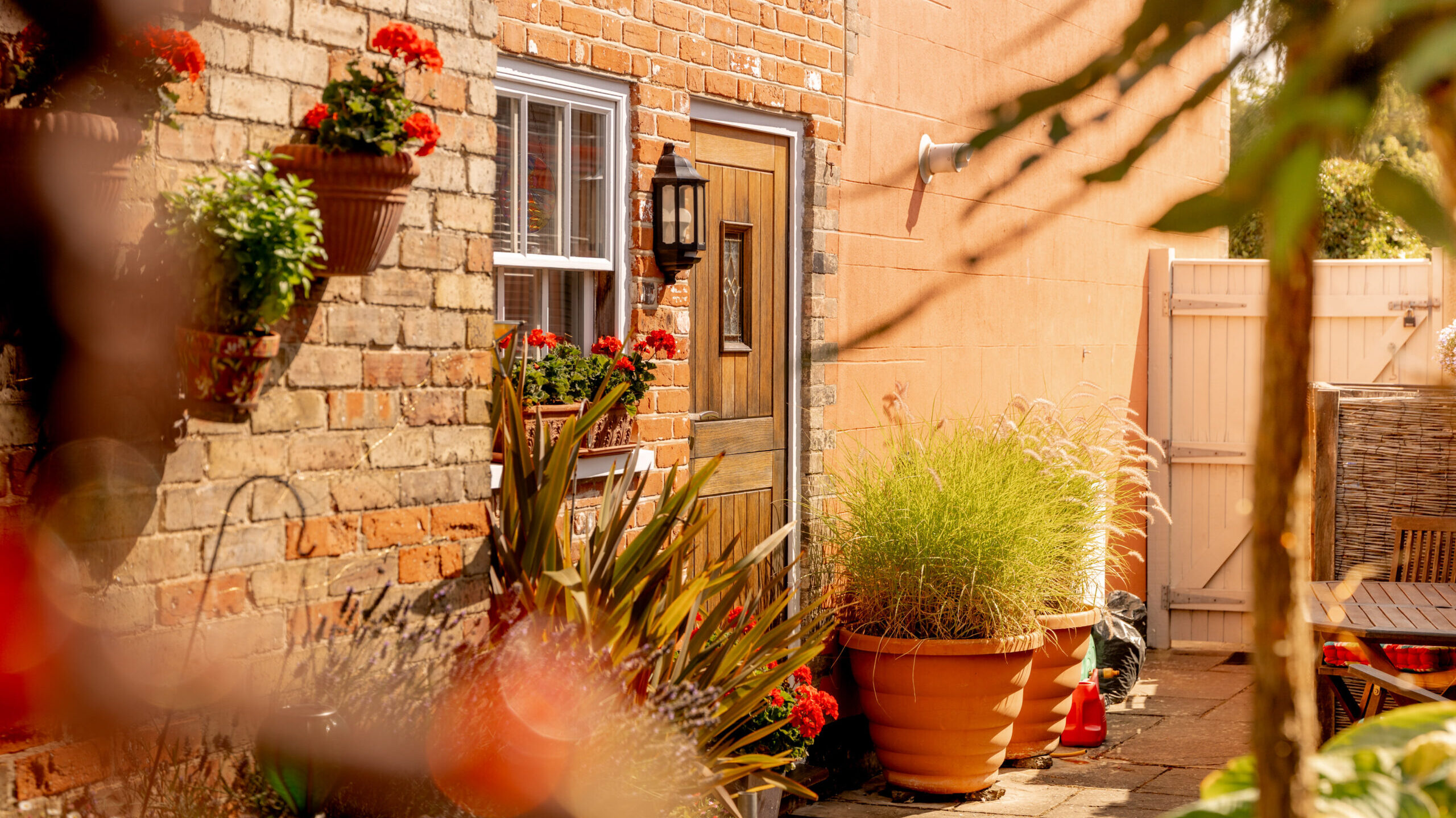 Sunny patio with brick walls, potted plants, red flowers, and a wooden door. Outdoor furniture is visible on the right, and a white fence encloses the cozy garden area.