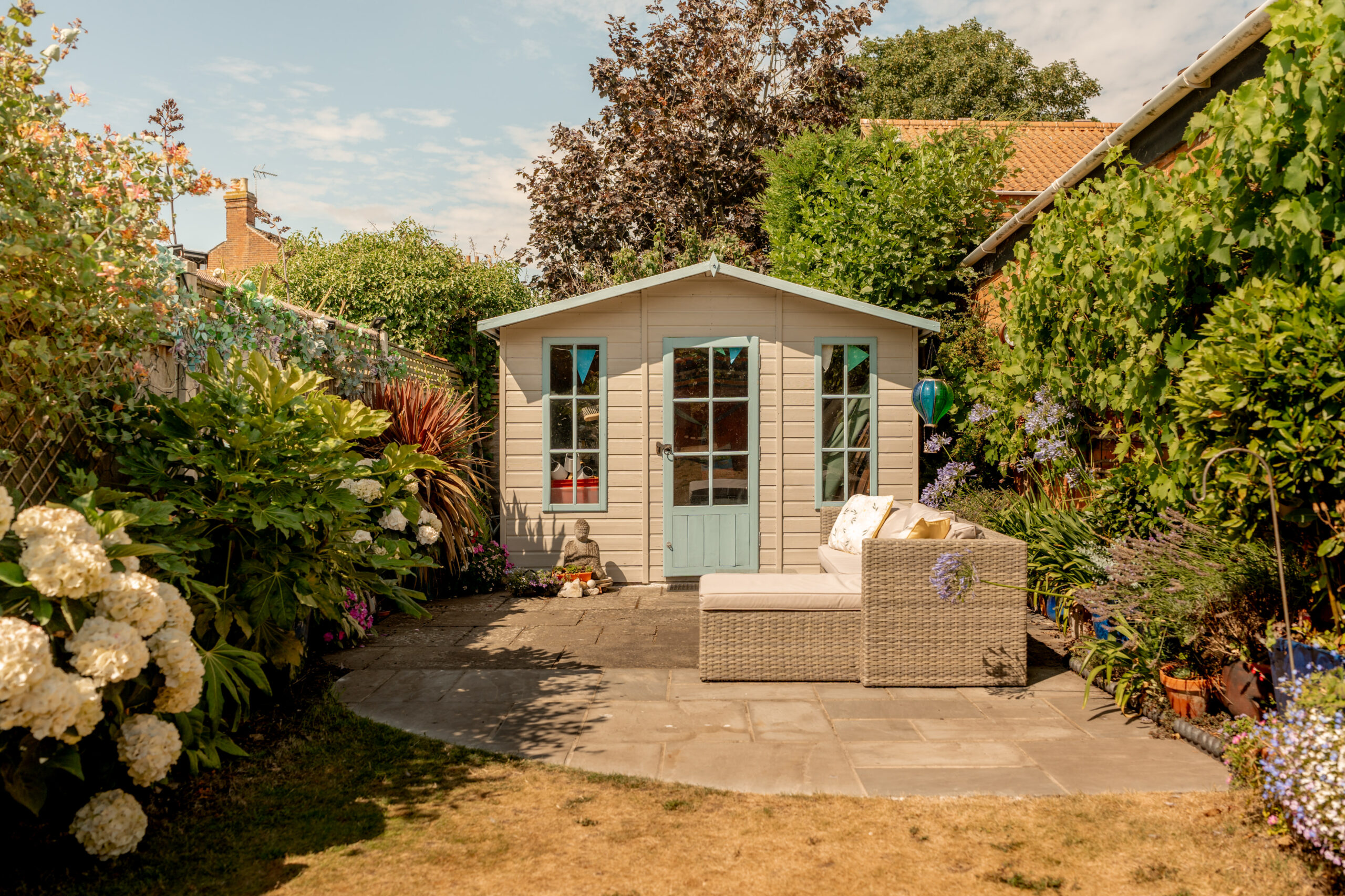 A small cream garden shed with blue accents sits at the end of a stone patio, surrounded by lush plants and flowers. A beige outdoor sofa with cushions is on the patio in front of the shed.