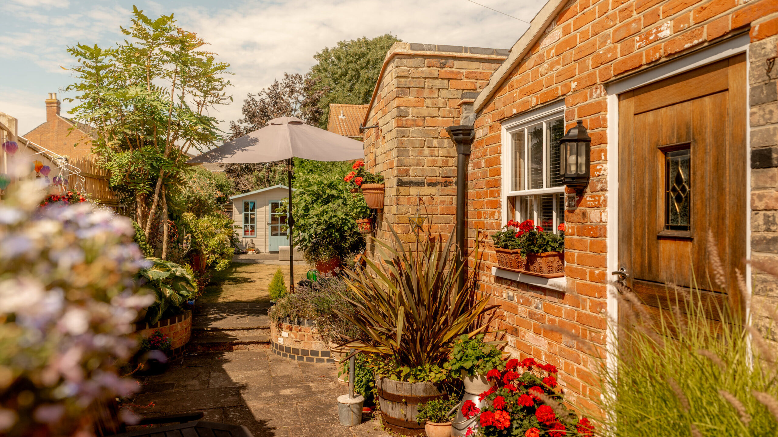 A charming brick house with a wooden door, potted flowers, and lush plants lining a sunlit garden path, leading to a small outbuilding under a large gray umbrella.