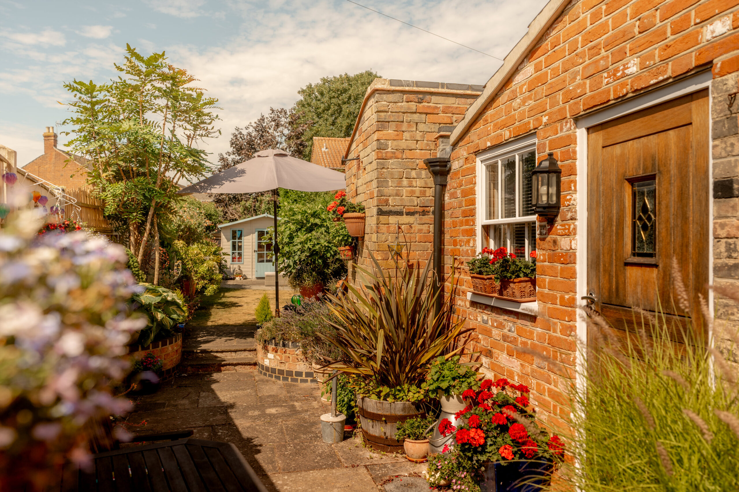A charming brick house with a wooden door, potted flowers, and lush plants lining a sunlit garden path, leading to a small outbuilding under a large gray umbrella.