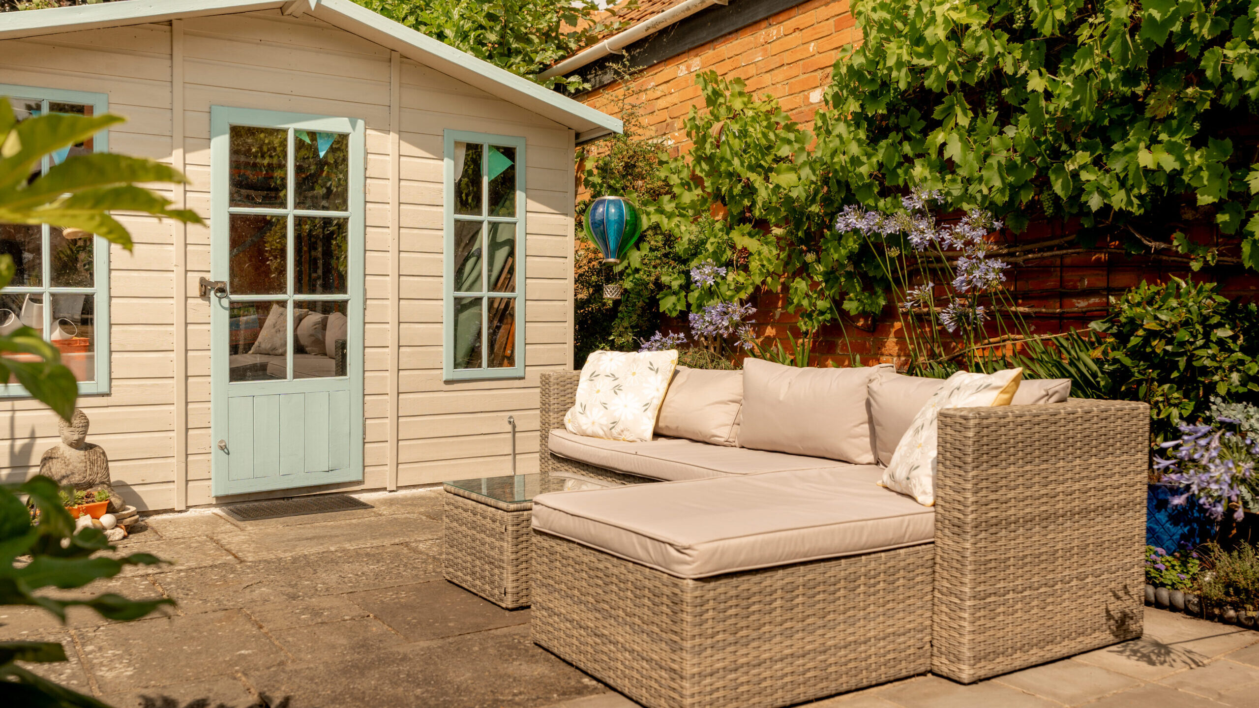 A cozy outdoor patio with a wicker sofa and table sits beside a light-colored garden shed surrounded by greenery and flowers, under a sunny sky.