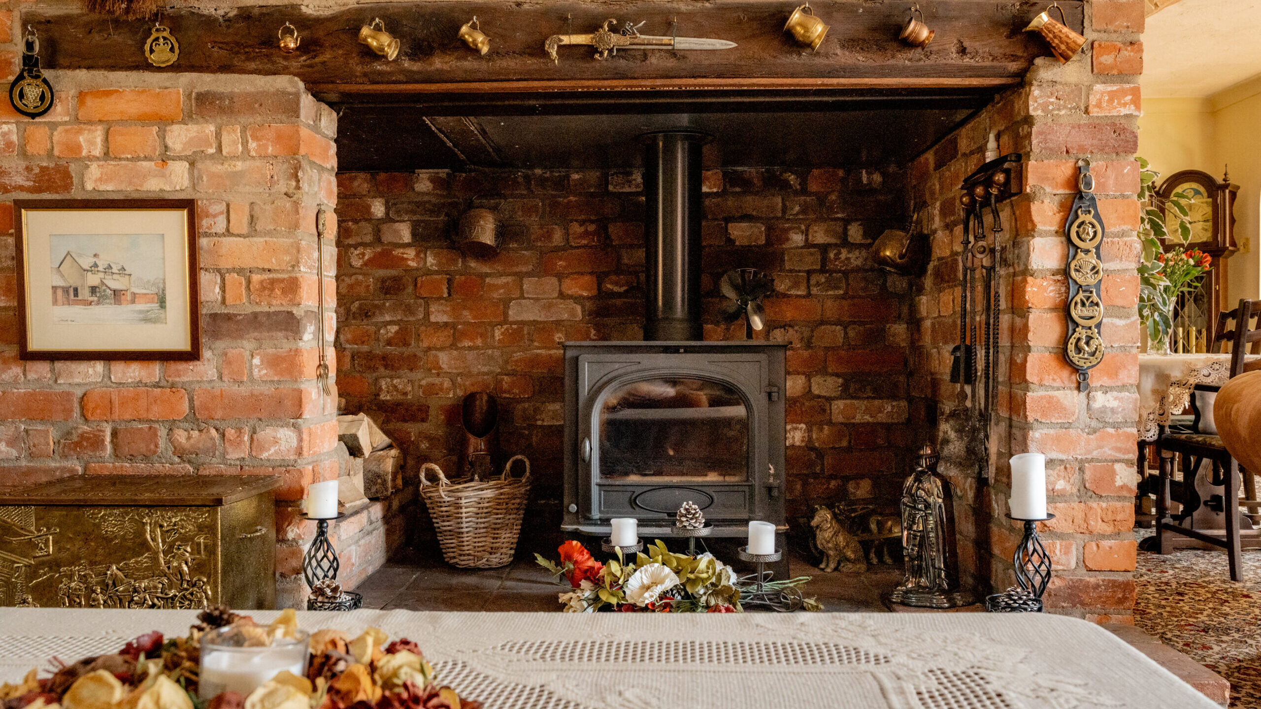 A cozy rustic living room with a brick fireplace, wood-burning stove, wicker basket, brass ornaments, candles, and a lace-covered table with a dried flower centerpiece in the foreground.