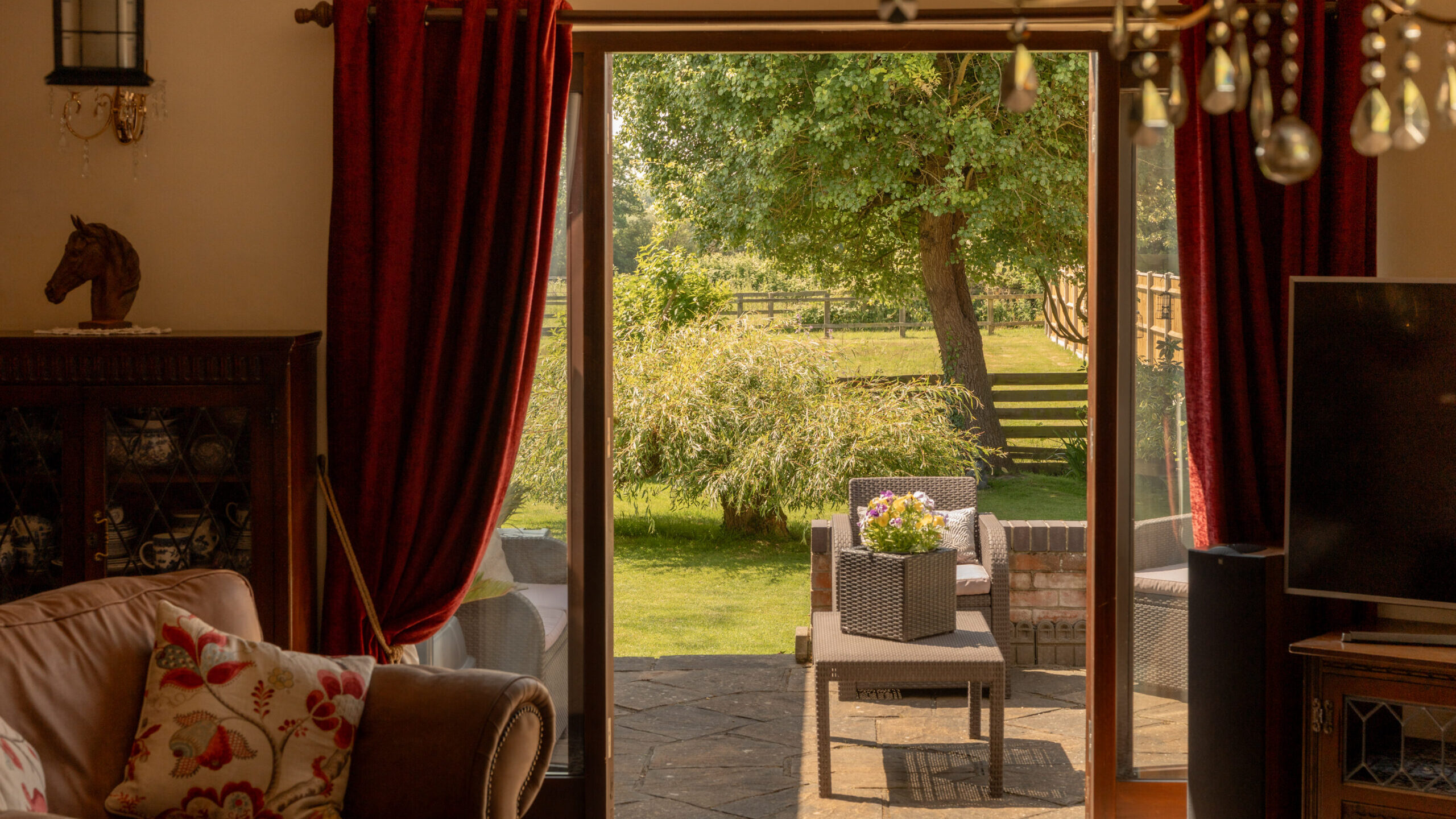 A cozy living room with red curtains and a brown sofa looks out through glass doors onto a sunny garden with patio furniture, green grass, and a large tree.
