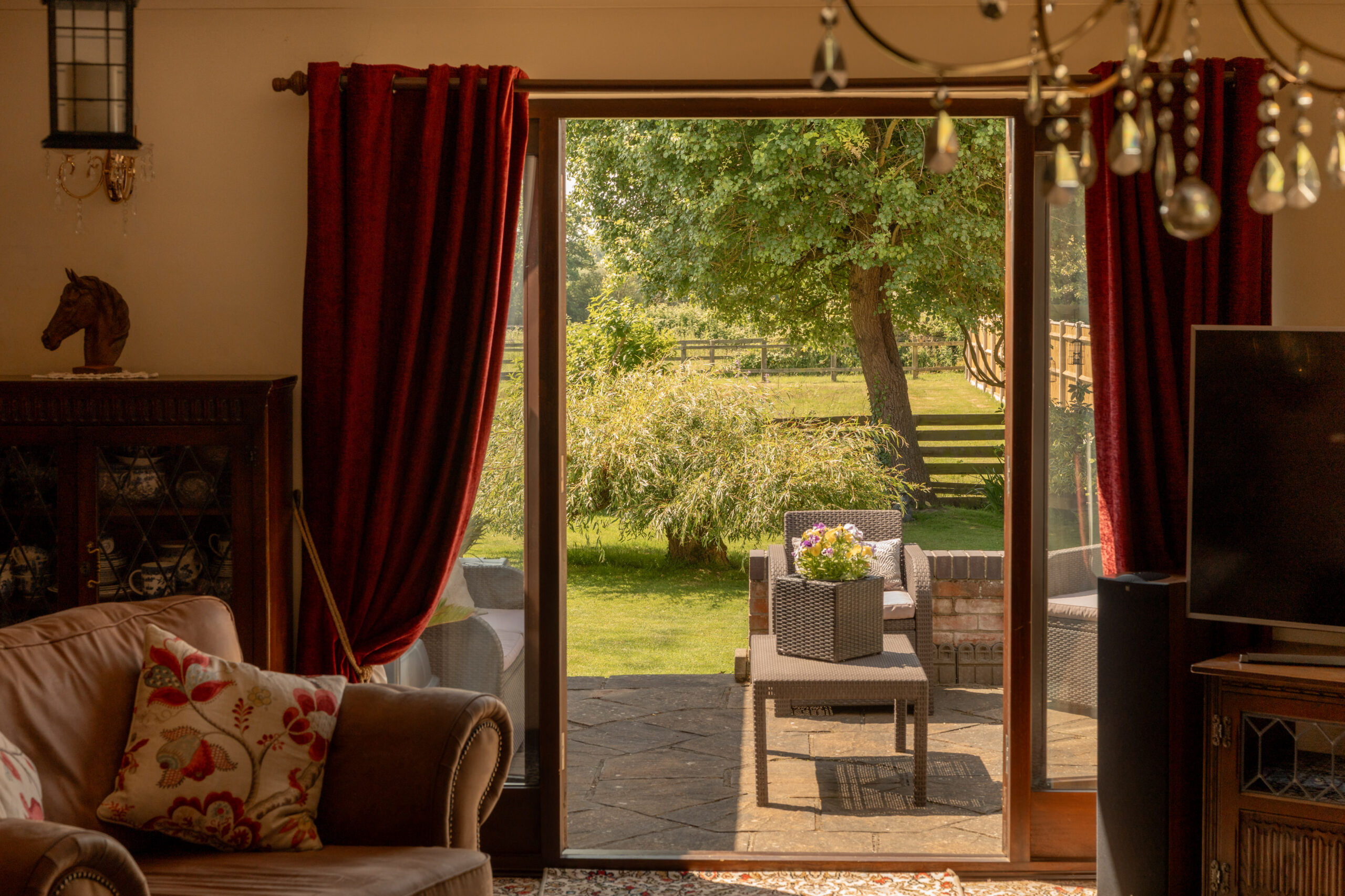 A cozy living room with red curtains and a brown sofa looks out through glass doors onto a sunny garden with patio furniture, green grass, and a large tree.