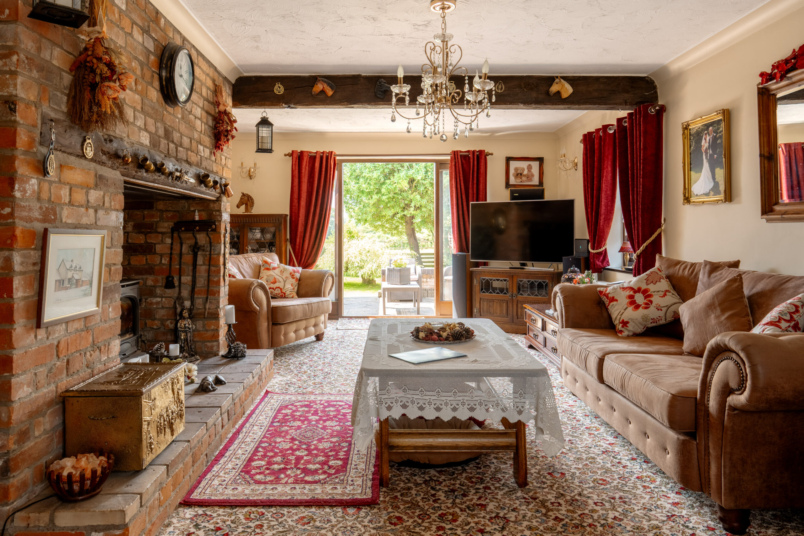 A cozy, traditionally decorated living room with a brick fireplace, brown sofas, patterned rug, ornate curtains, a chandelier, and a large window overlooking a green garden. A TV and framed art complete the space.