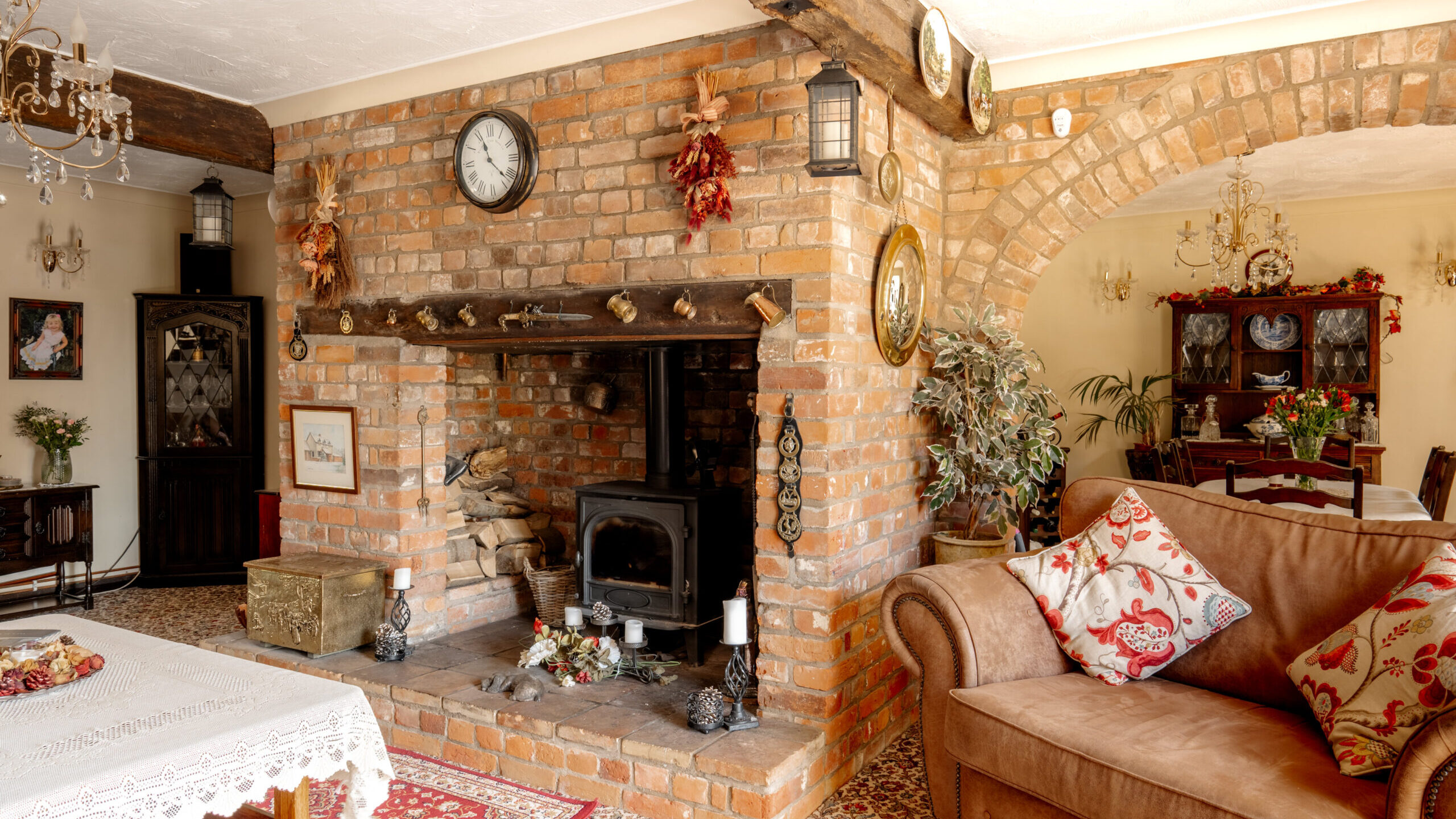 A cozy living room with a brick fireplace and wood stove, decorated with candles, plants, and a clock. There’s a brown sofa with patterned pillows and a dining area with a table and chairs in the background.