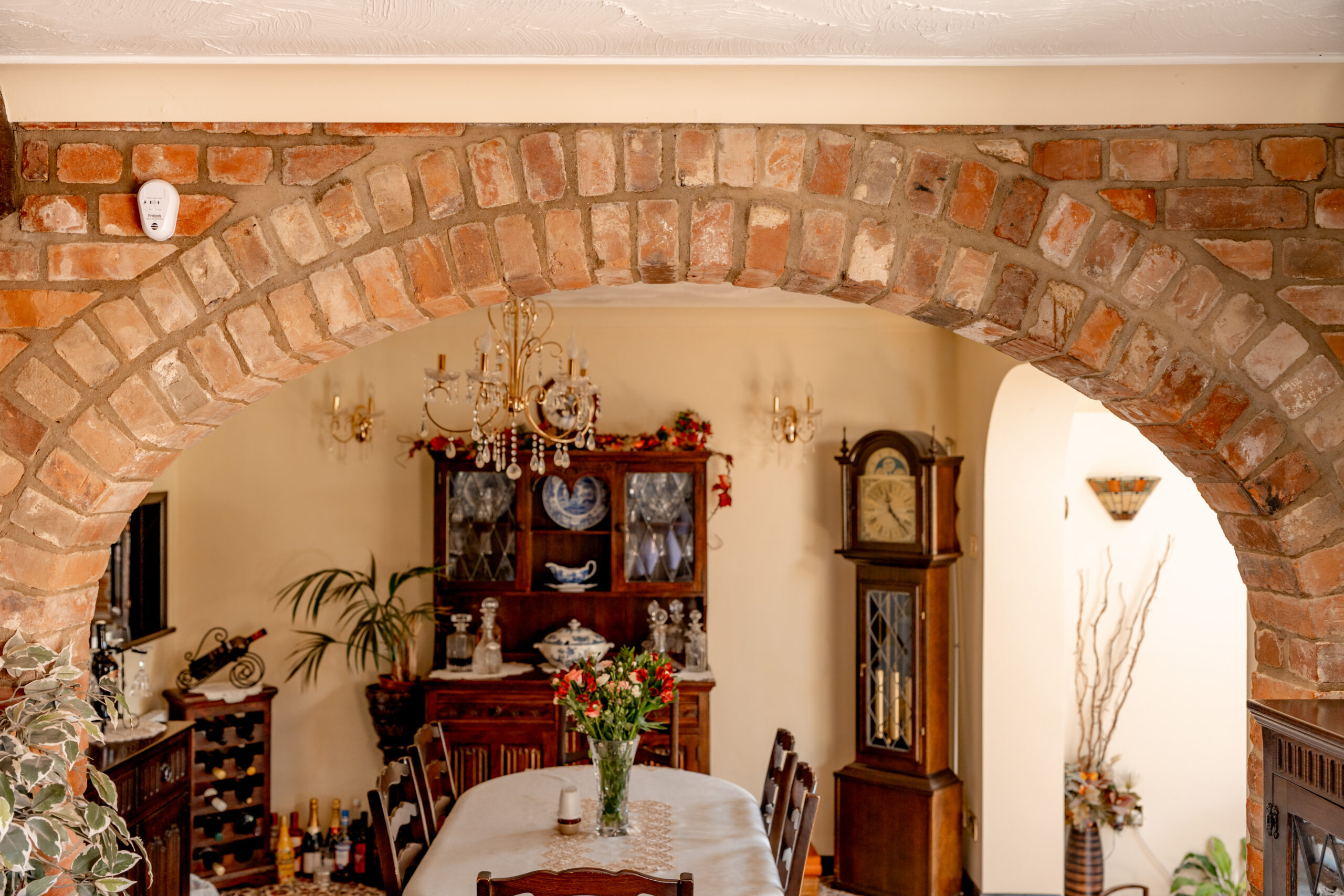 A cozy dining room with a brick archway, wooden furniture, a grandfather clock, a chandelier, and a table set with flowers and candles. Decorative plants and a china cabinet with glassware are in the background.