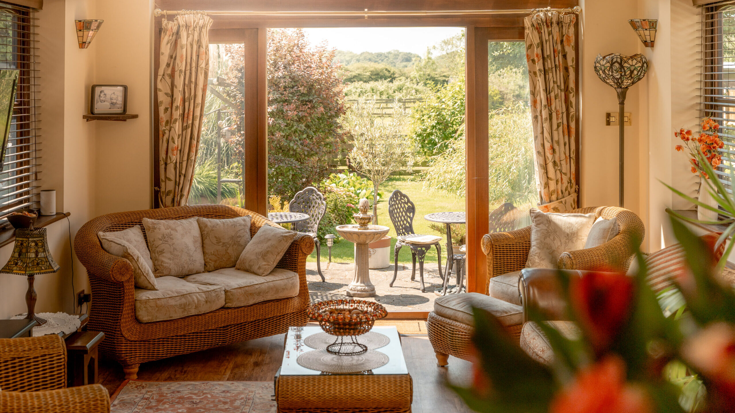 A cozy sunlit living room with wicker furniture faces large glass doors that open to a garden patio with a small table and two chairs, surrounded by lush greenery and trees.