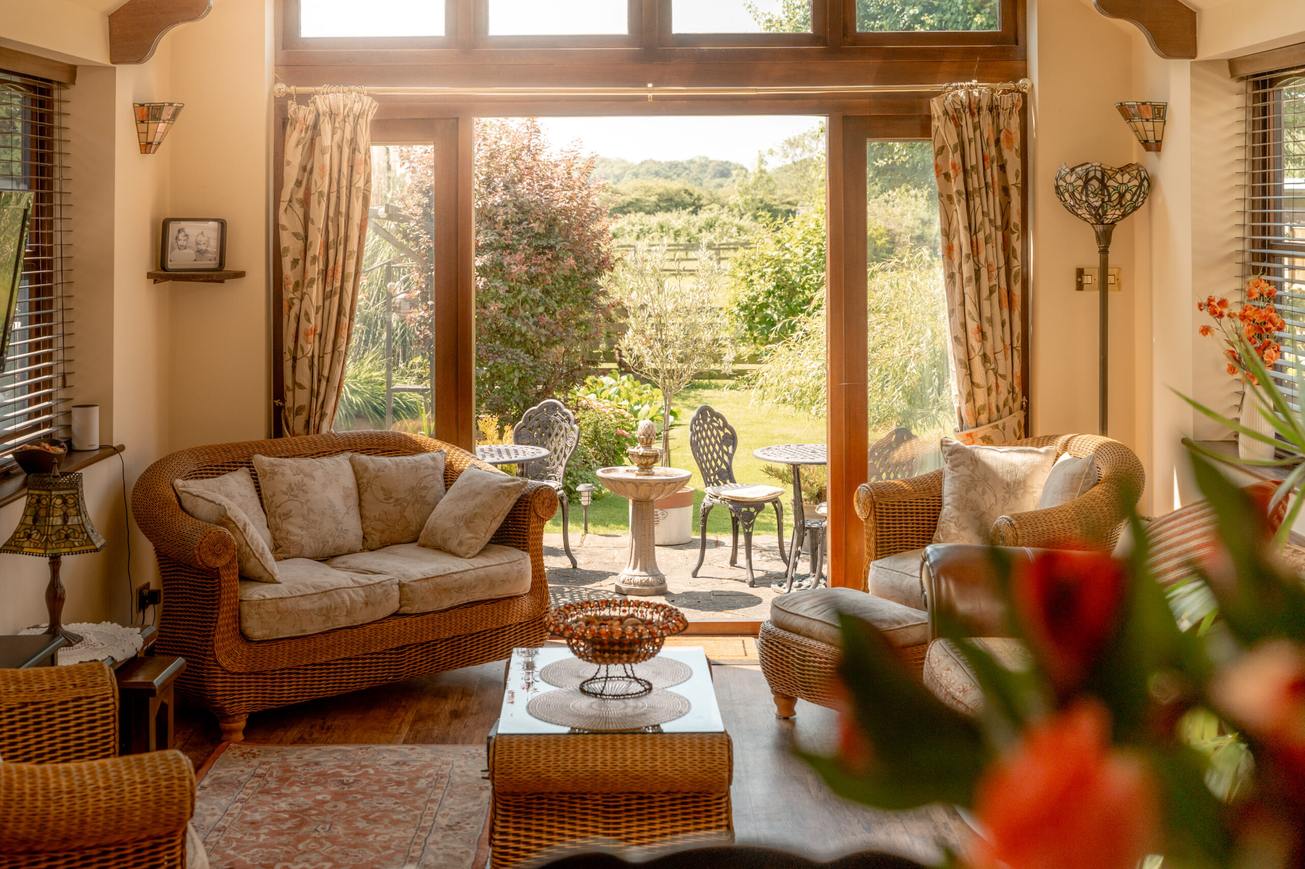 A cozy sunlit living room with wicker furniture faces large glass doors that open to a garden patio with a small table and two chairs, surrounded by lush greenery and trees.