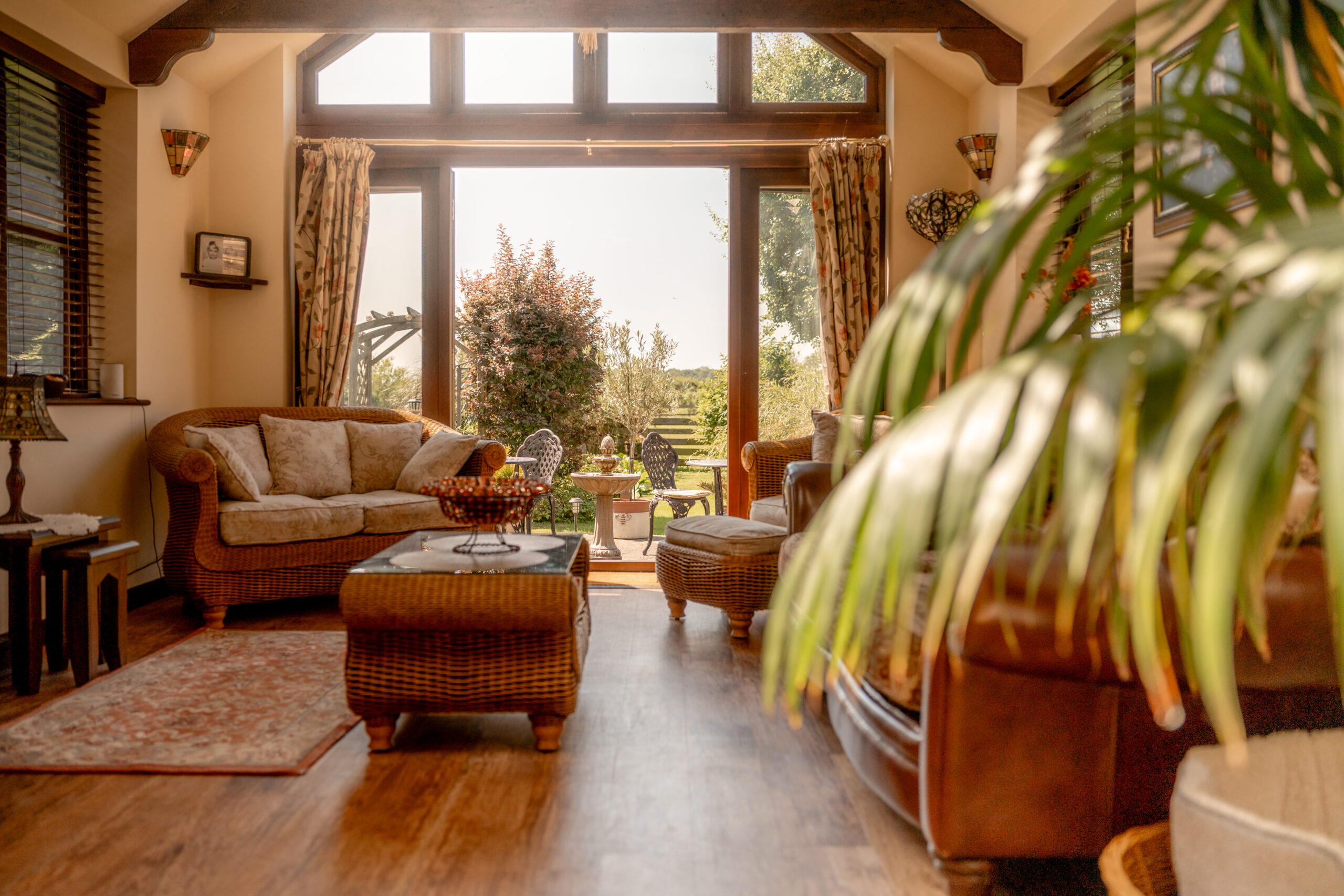 A cozy, sunlit living room with wicker furniture, a sofa, and plants, featuring large windows and glass doors that open to a garden patio with chairs and greenery outside.
