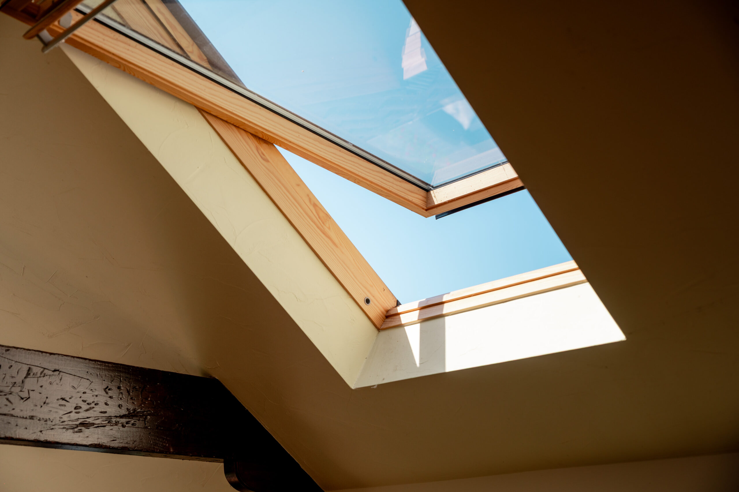 A skylight window set in a slanted ceiling allows sunlight to stream into the room, with a clear blue sky visible through the glass. A wooden beam and textured walls frame the window opening.