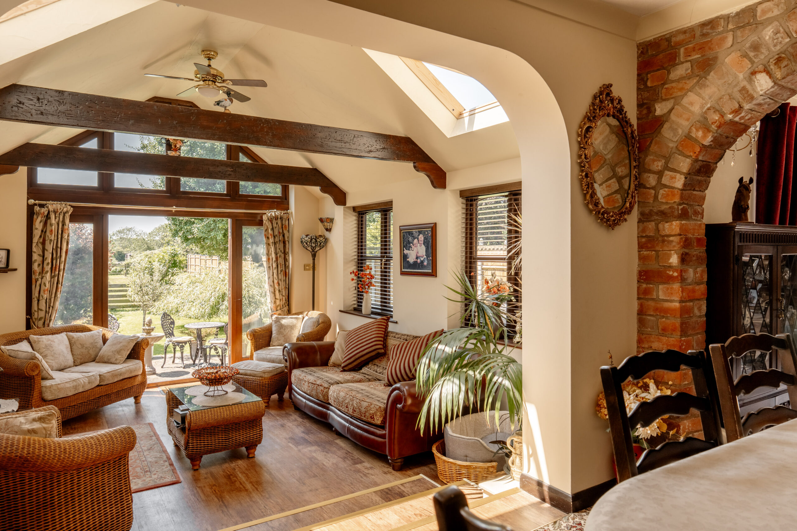 A cozy, sunlit living room with wicker and upholstered furniture, large windows and glass doors opening to a garden, and rustic wooden beams on the ceiling. There is a brick archway leading to a dining area.