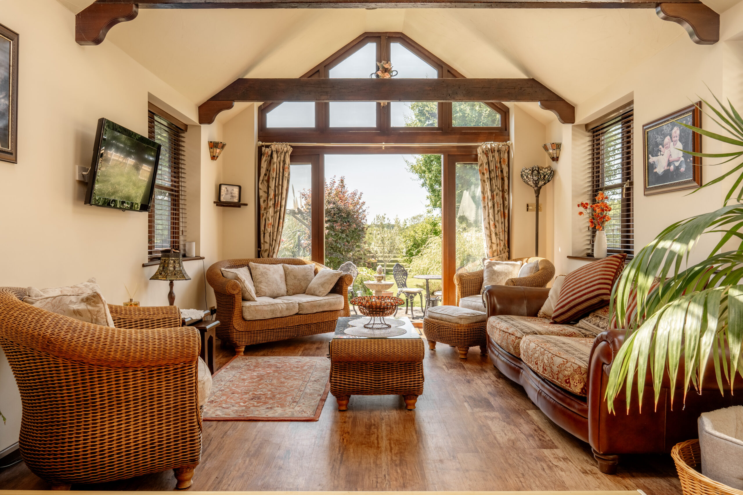 Bright sunroom with wicker and upholstered furniture, wooden floors, plants, and decor. Large windows and glass doors open to a sunny garden, letting in natural light. Exposed beams and cozy seating create a welcoming atmosphere.