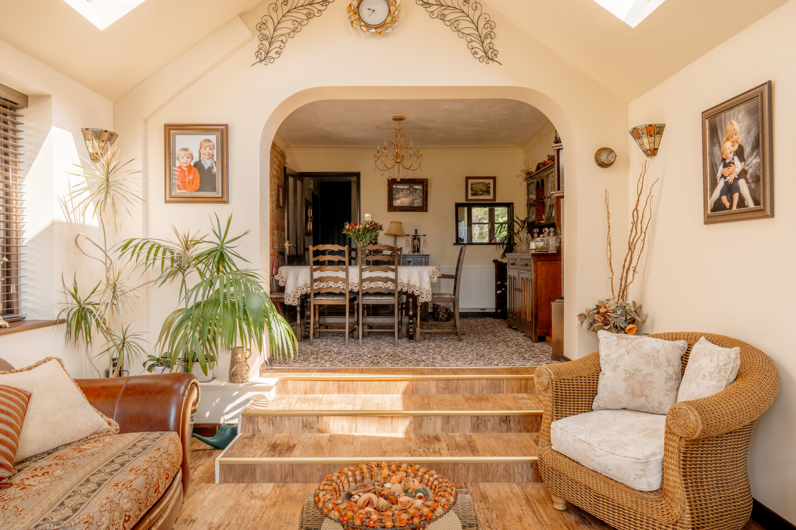 Bright, cozy living room with a wicker chair, leather sofa, potted plants, and steps leading to a dining area. Family photos, wall art, and a clock decorate the cream-colored walls. Sunlight streams in through skylights.