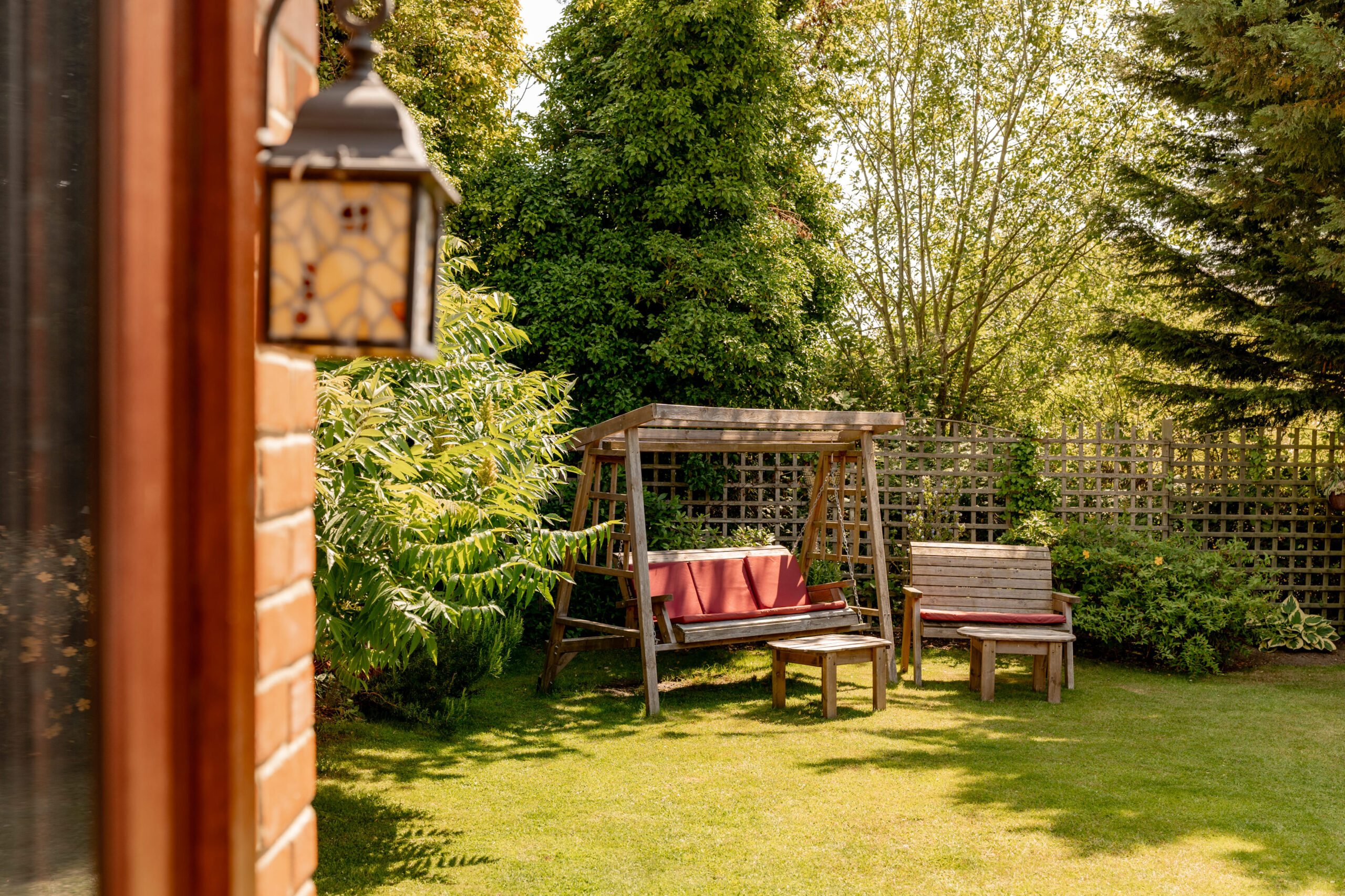 A sunlit garden features a wooden swing with red cushions, a wooden bench, and a small table on green grass, surrounded by trees and shrubs. A lantern hangs on the left near a brick wall.