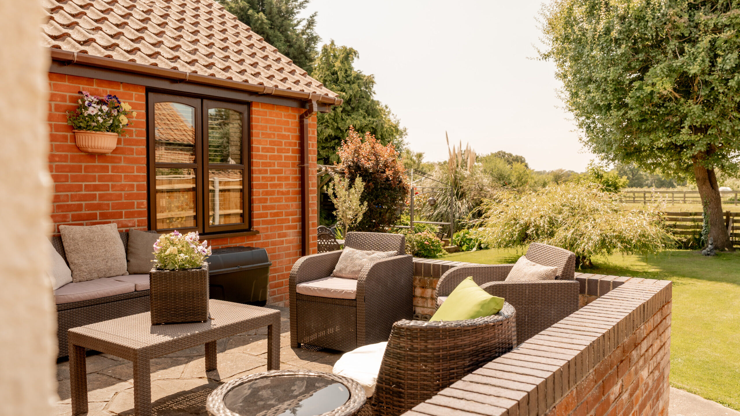 Sunny patio with wicker chairs, a coffee table, and cushions, set beside a brick house. Green lawn, potted plants, trees, and a wooden fence can be seen in the background.