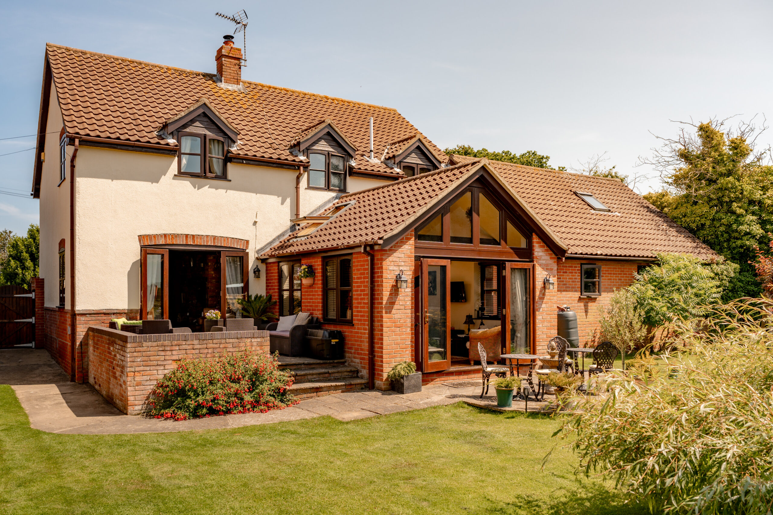 A two-story house with white walls and red roof tiles, featuring large windows, a patio with outdoor furniture, and a well-kept garden with green grass and plants in the backyard.