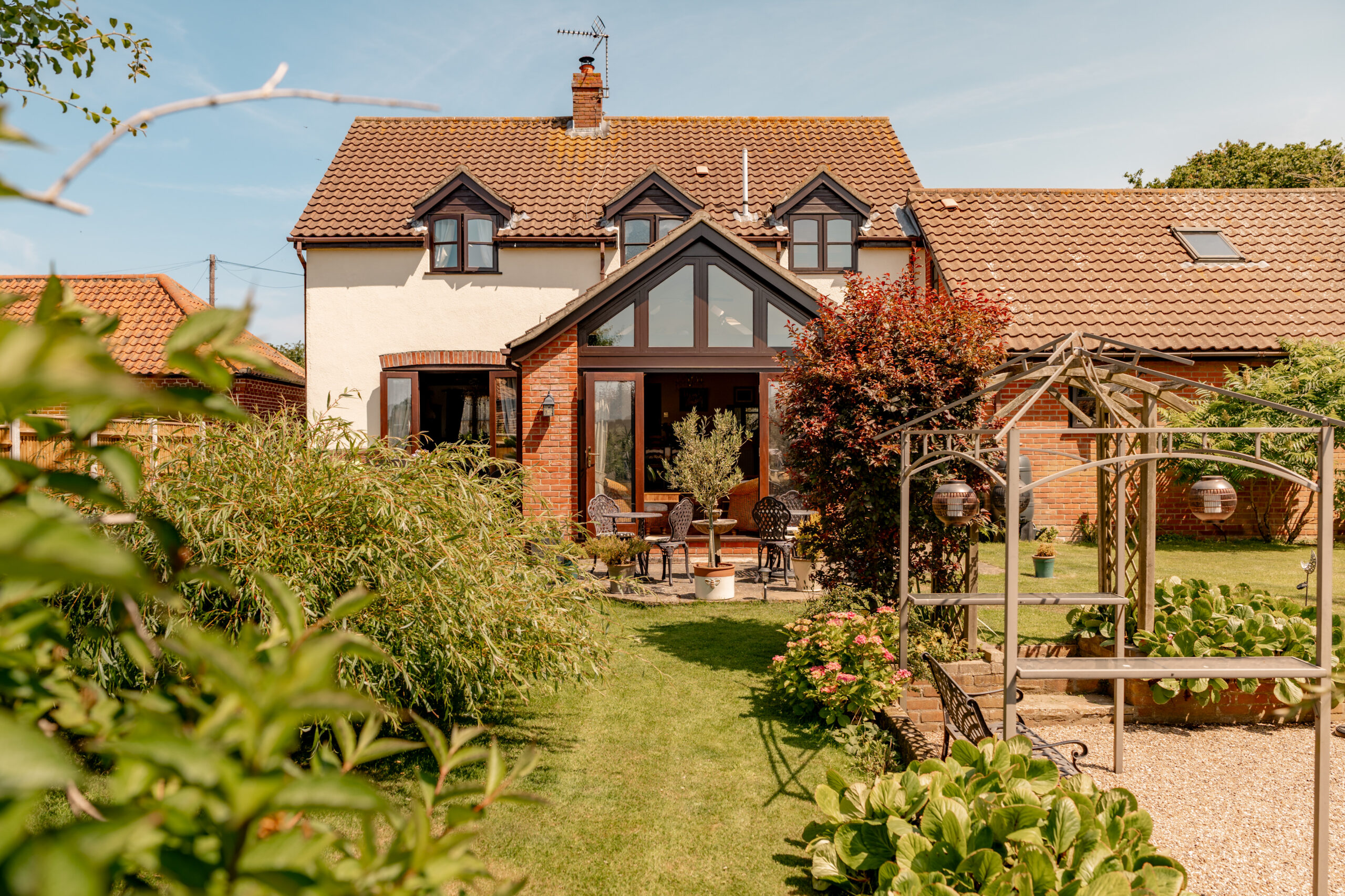 A charming two-story house with a red-tiled roof, large windows, and a lush garden featuring patio furniture, greenery, and a wooden pergola on a sunny day.
