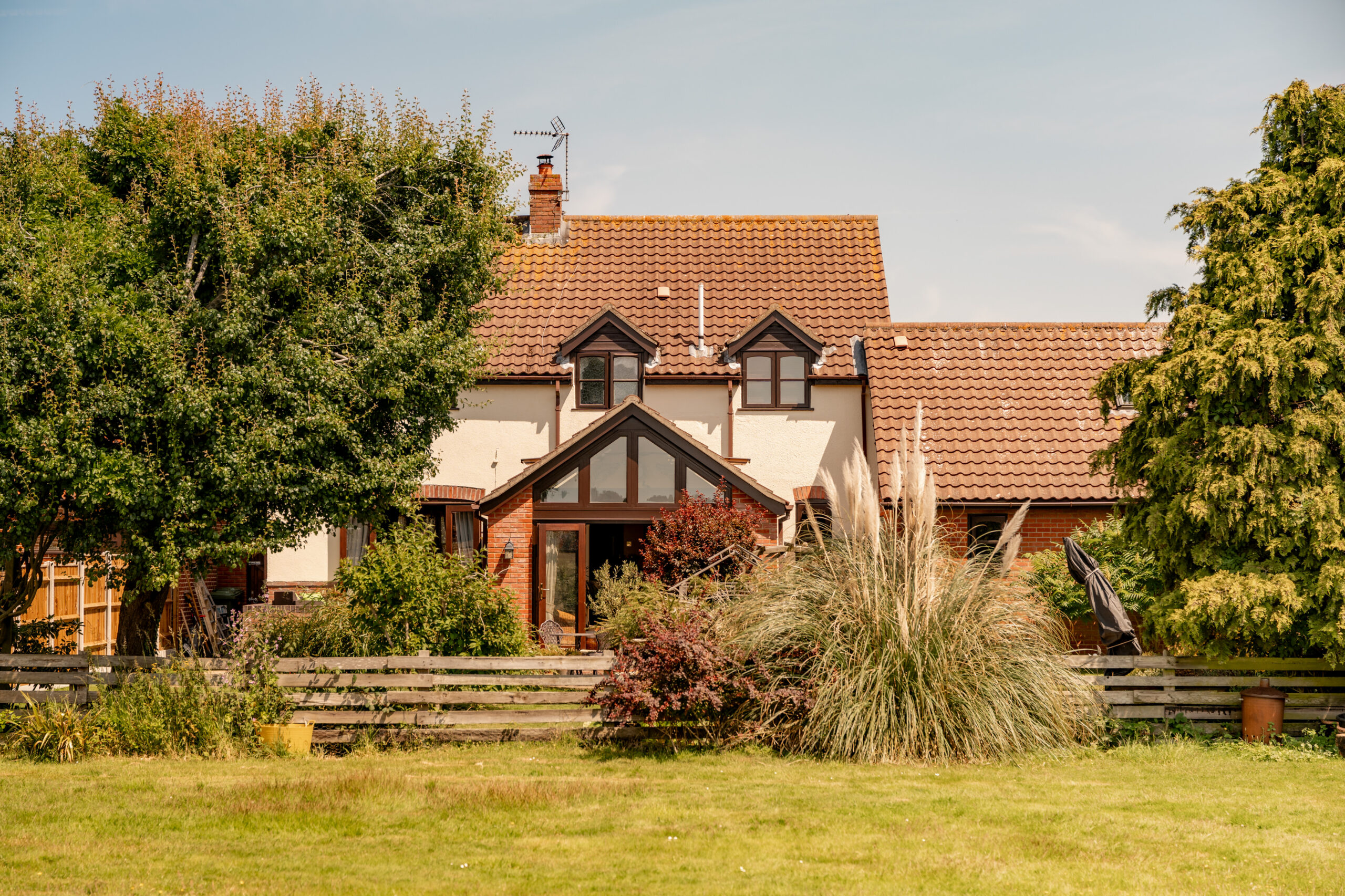 A two-story house with tan walls and a red-tiled roof, surrounded by green trees, shrubs, and a grassy lawn, seen on a sunny day. A wooden fence runs in front of the property.