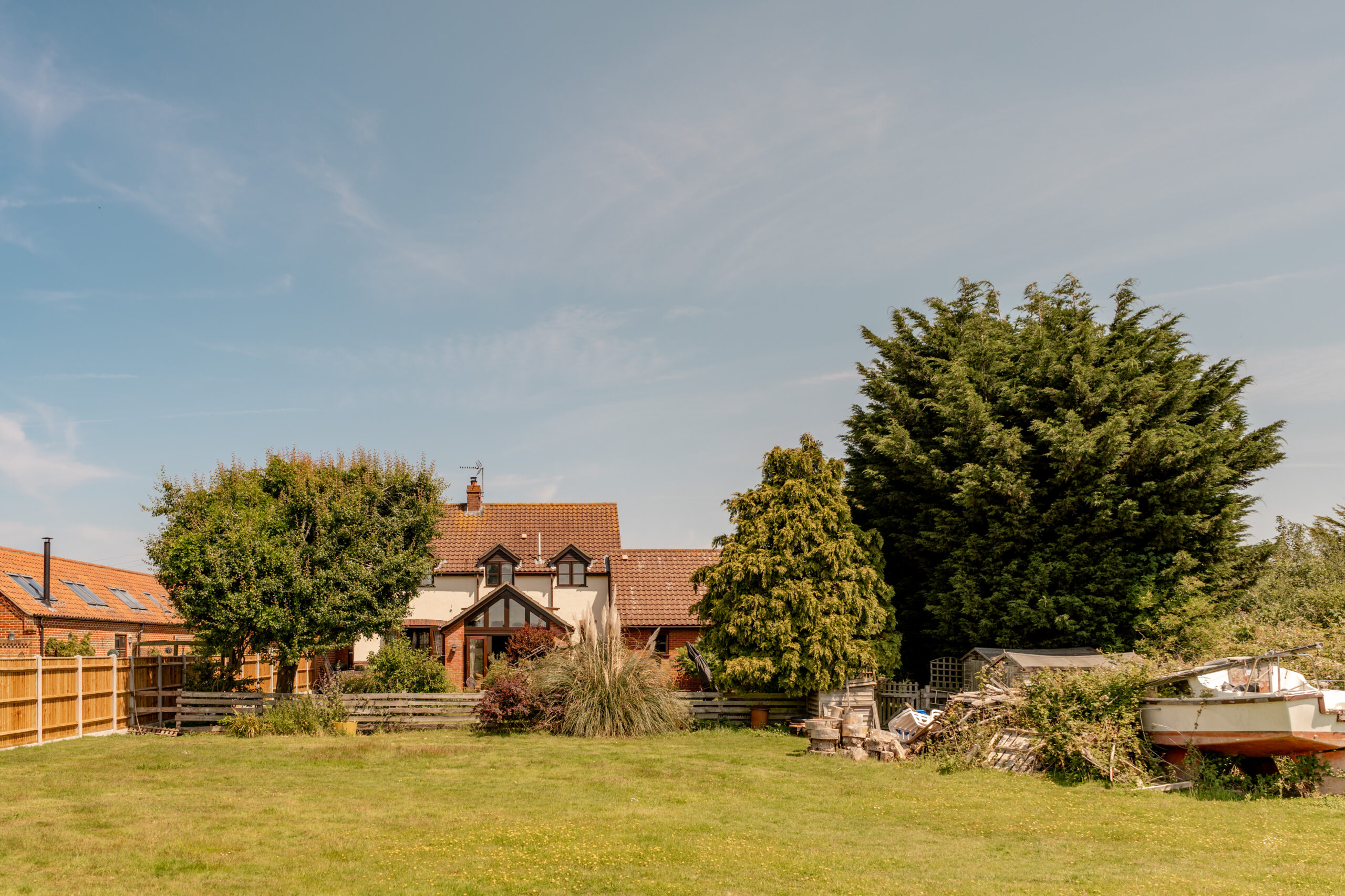 A house with a red-tiled roof surrounded by trees and bushes, with a large grassy yard in front. A small boat sits on the right side near a pile of wood, and the sky is clear and blue.
