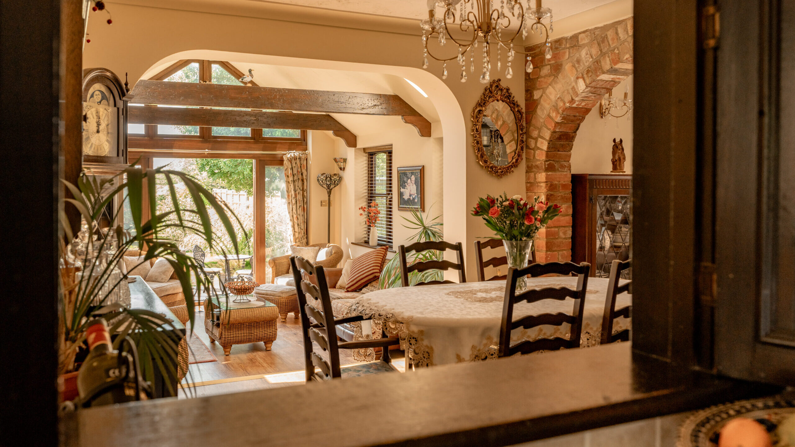 A warm, inviting dining and living area viewed through a kitchen window, featuring a round table with flowers, wooden chairs, vintage décor, and sunlight streaming in through large windows.