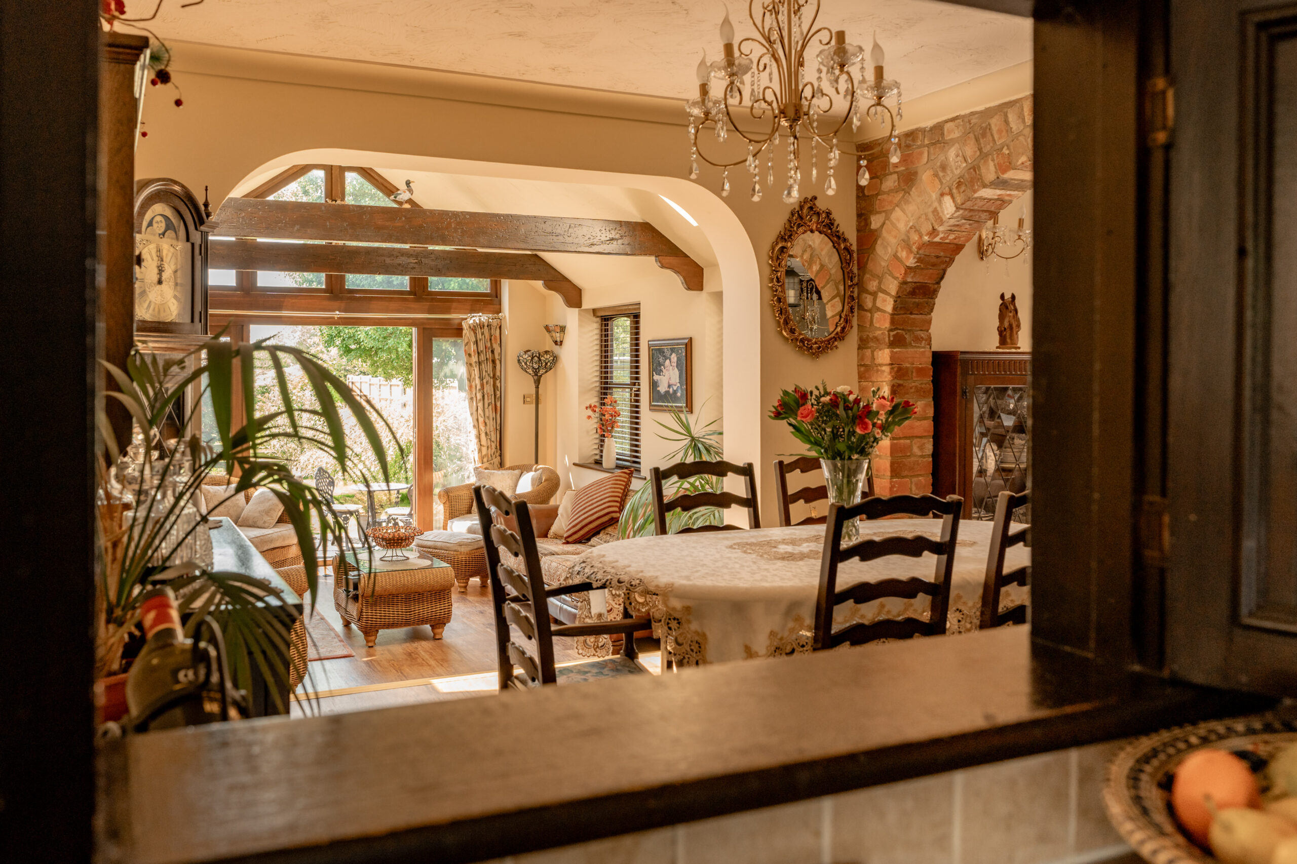 A warm, inviting dining and living area viewed through a kitchen window, featuring a round table with flowers, wooden chairs, vintage décor, and sunlight streaming in through large windows.