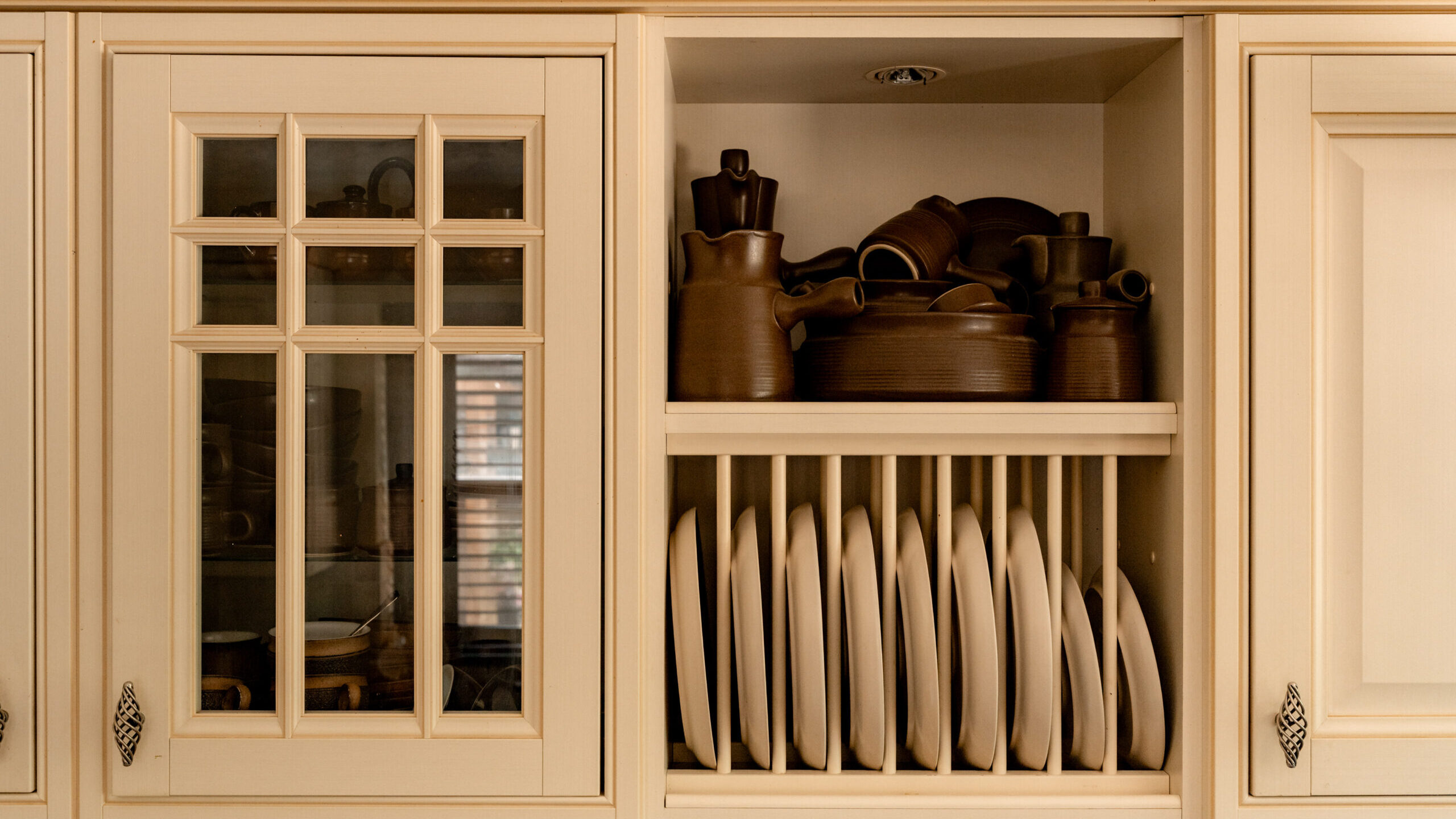 A kitchen cabinet with an open section displaying brown pottery and dishes on the top shelf and neatly stacked white plates in a vertical rack on the bottom shelf. The surrounding cabinets are cream-colored with glass and solid doors.
