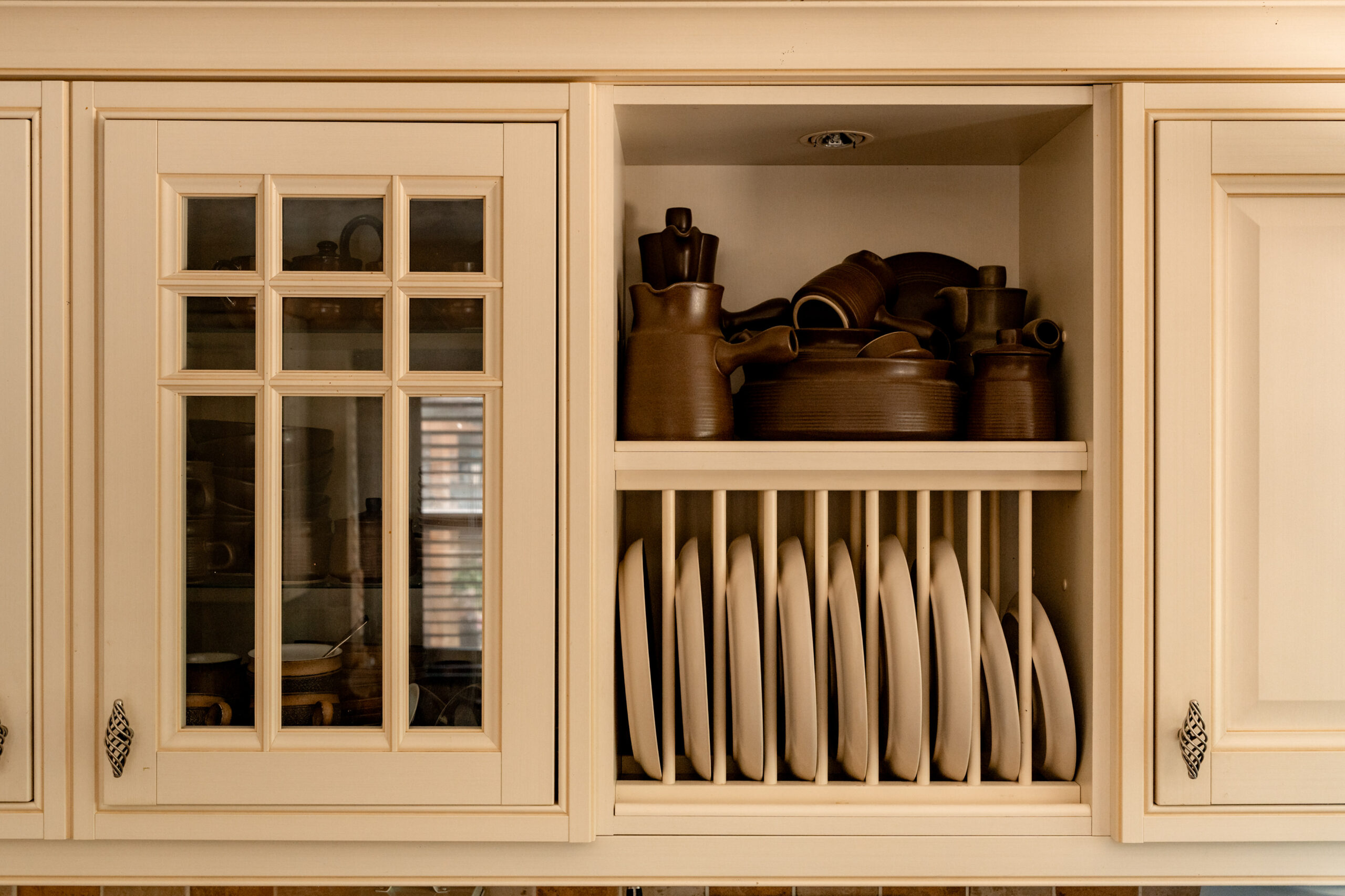 A kitchen cabinet with an open section displaying brown pottery and dishes on the top shelf and neatly stacked white plates in a vertical rack on the bottom shelf. The surrounding cabinets are cream-colored with glass and solid doors.