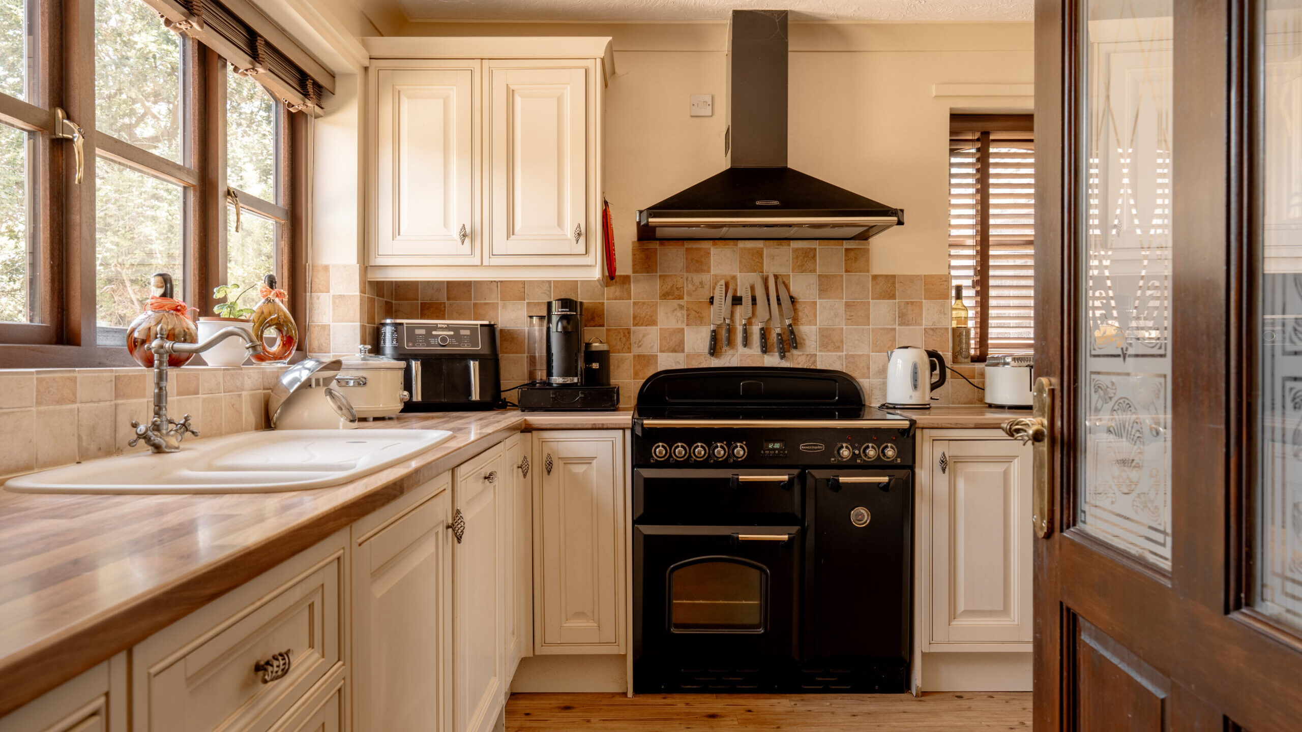 A cozy kitchen with cream cabinets, a wooden countertop, a black stove with an oven, a toaster, kettle, and coffee maker. Sunlight streams through the windows, highlighting the tiled backsplash and wooden floor.