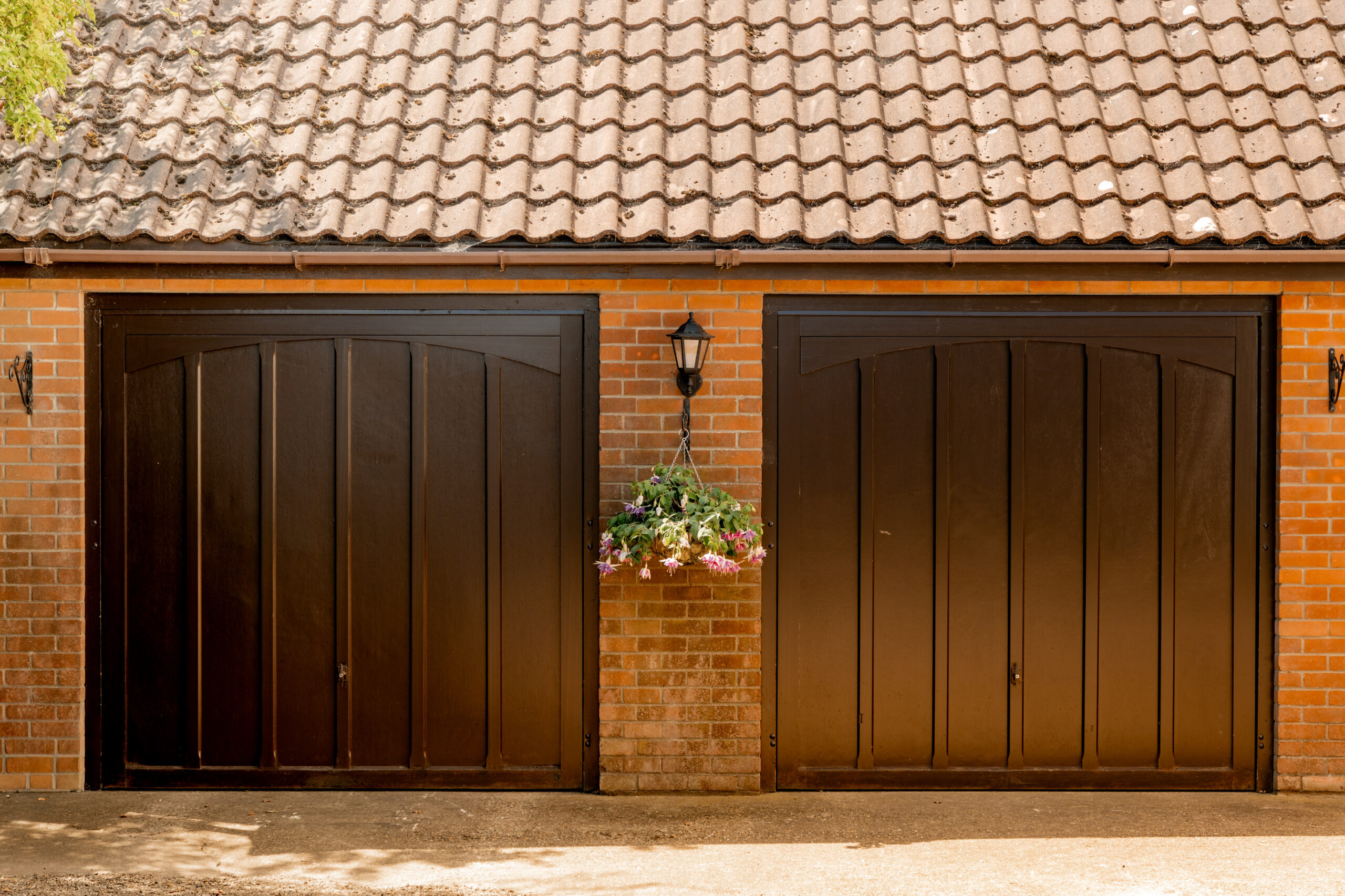 Two dark brown wooden garage doors on a brick building. A hanging basket with purple and white flowers is suspended between the doors beneath an outdoor wall lantern. The roof is covered with brown tiles.