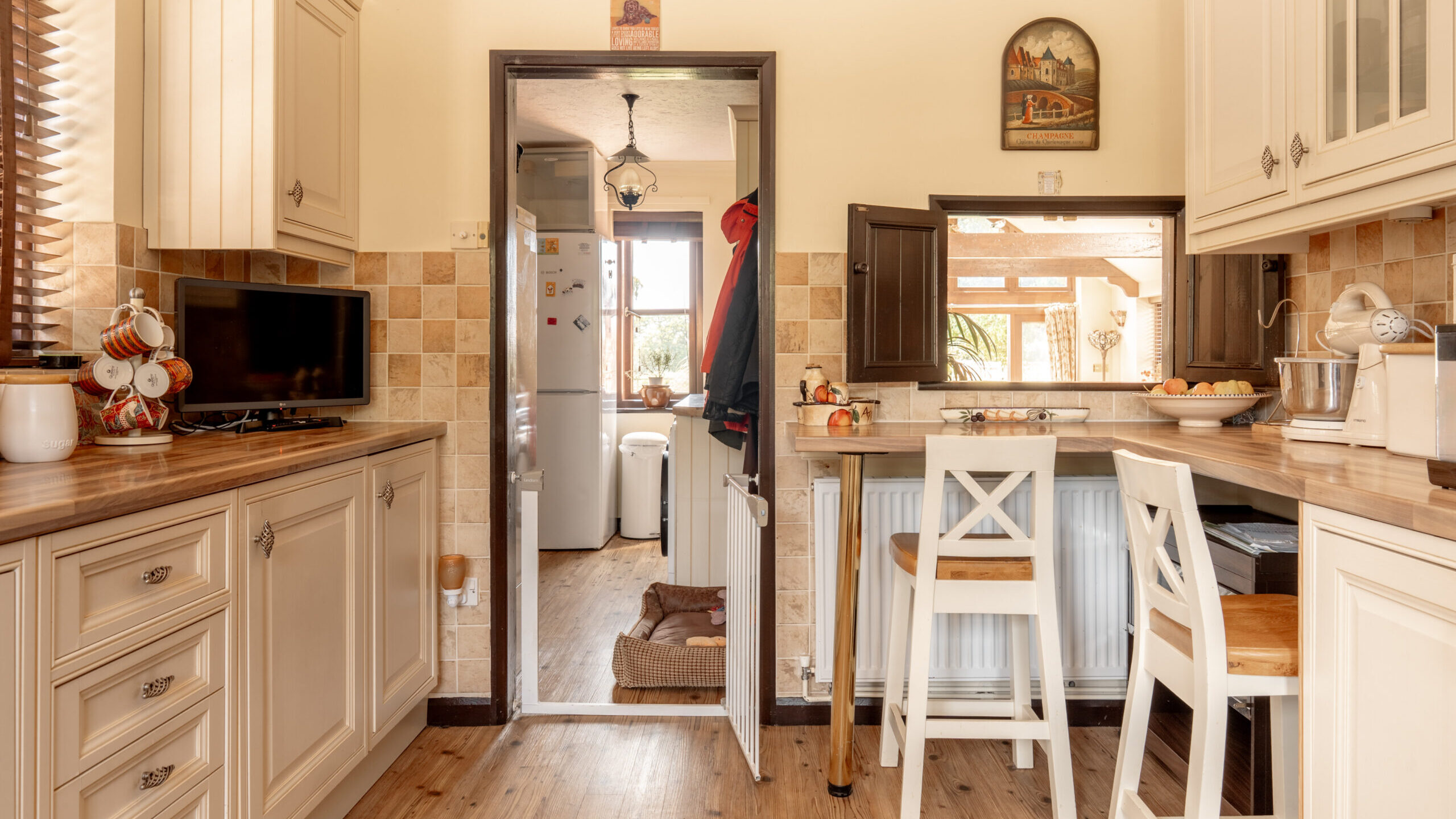 A cozy kitchen with cream cabinets, wood countertops, and tiled backsplash. There are two white chairs at a breakfast bar, a small TV, and a window looking out to another room with a dog bed on the floor.