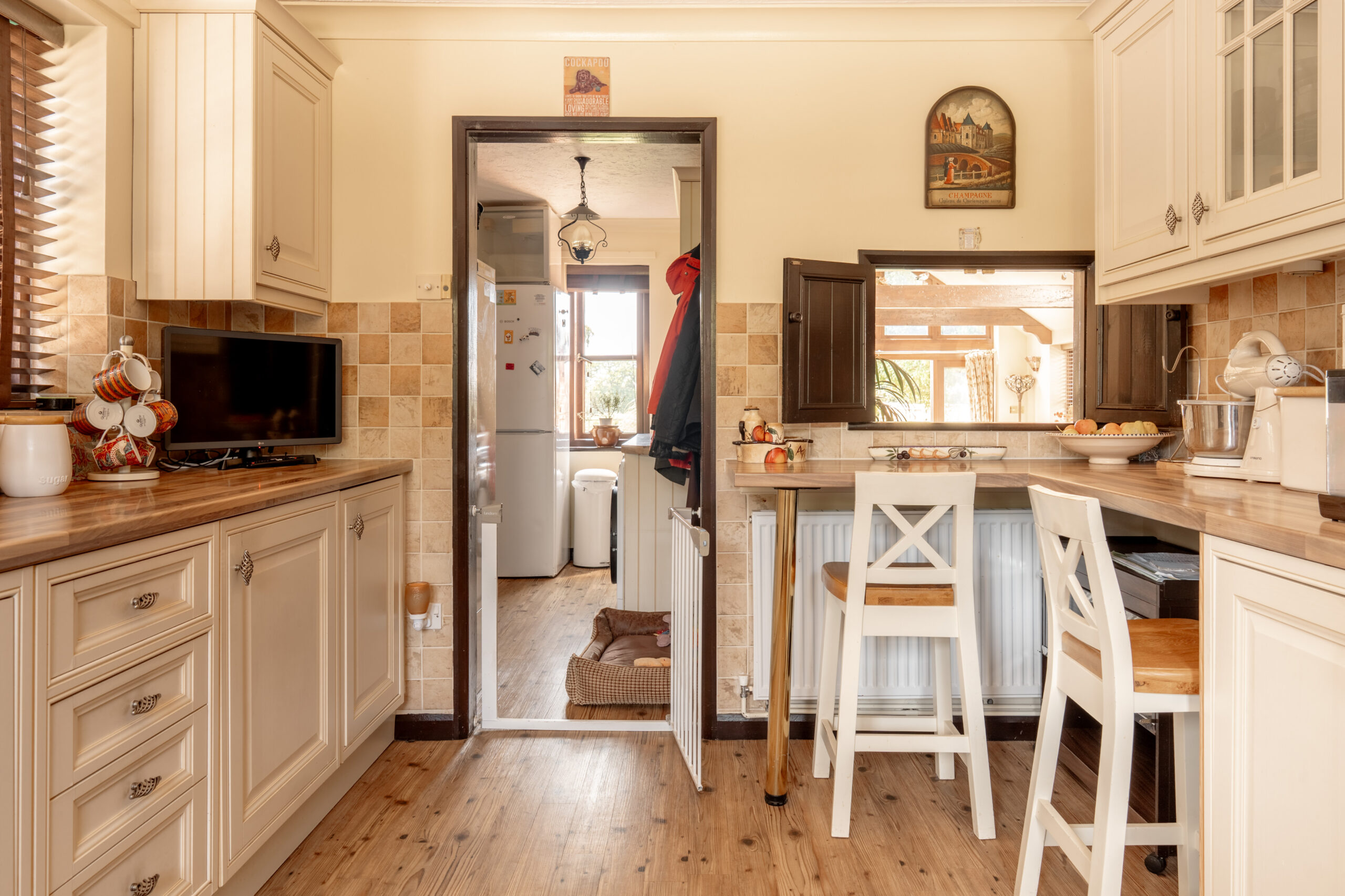 A cozy kitchen with cream cabinets, wood countertops, and tiled backsplash. There are two white chairs at a breakfast bar, a small TV, and a window looking out to another room with a dog bed on the floor.