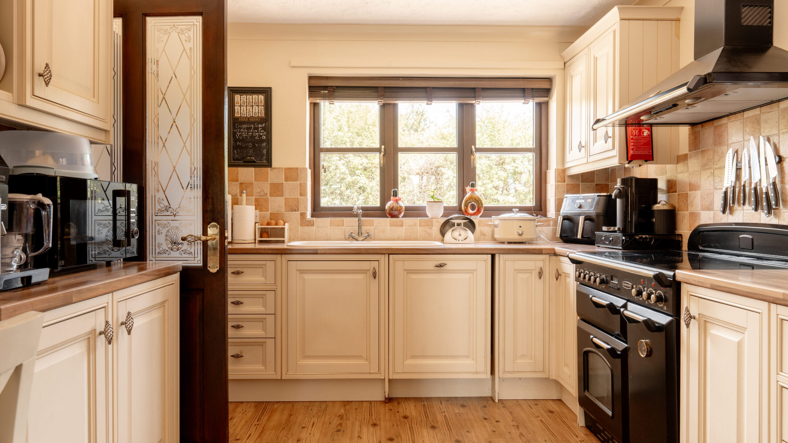 A cozy, traditional kitchen with cream-colored cabinets, a black oven and stove, small appliances on the counters, a wooden floor, and sunlight streaming through a window above the sink.