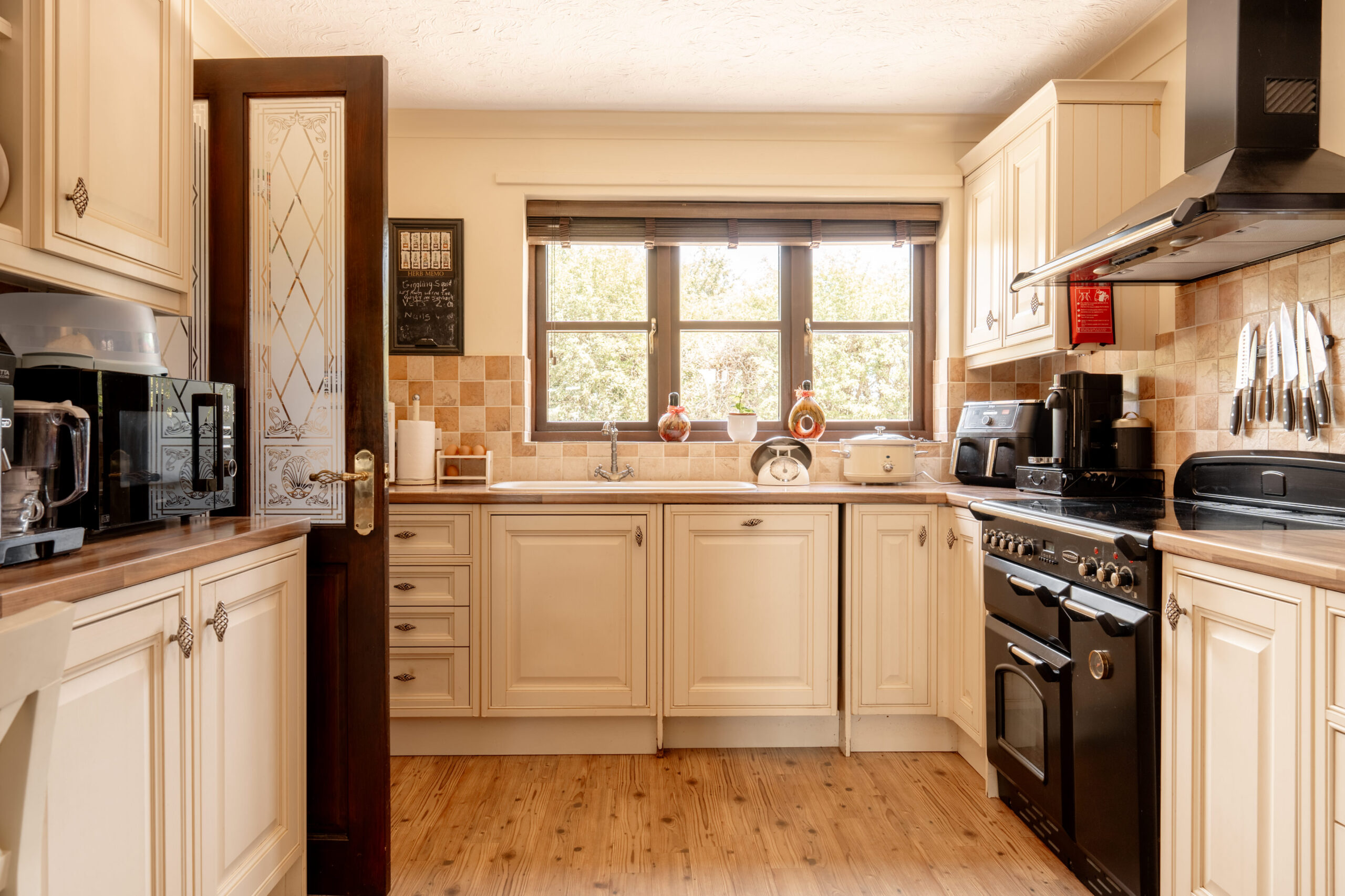 A cozy, traditional kitchen with cream-colored cabinets, a black oven and stove, small appliances on the counters, a wooden floor, and sunlight streaming through a window above the sink.