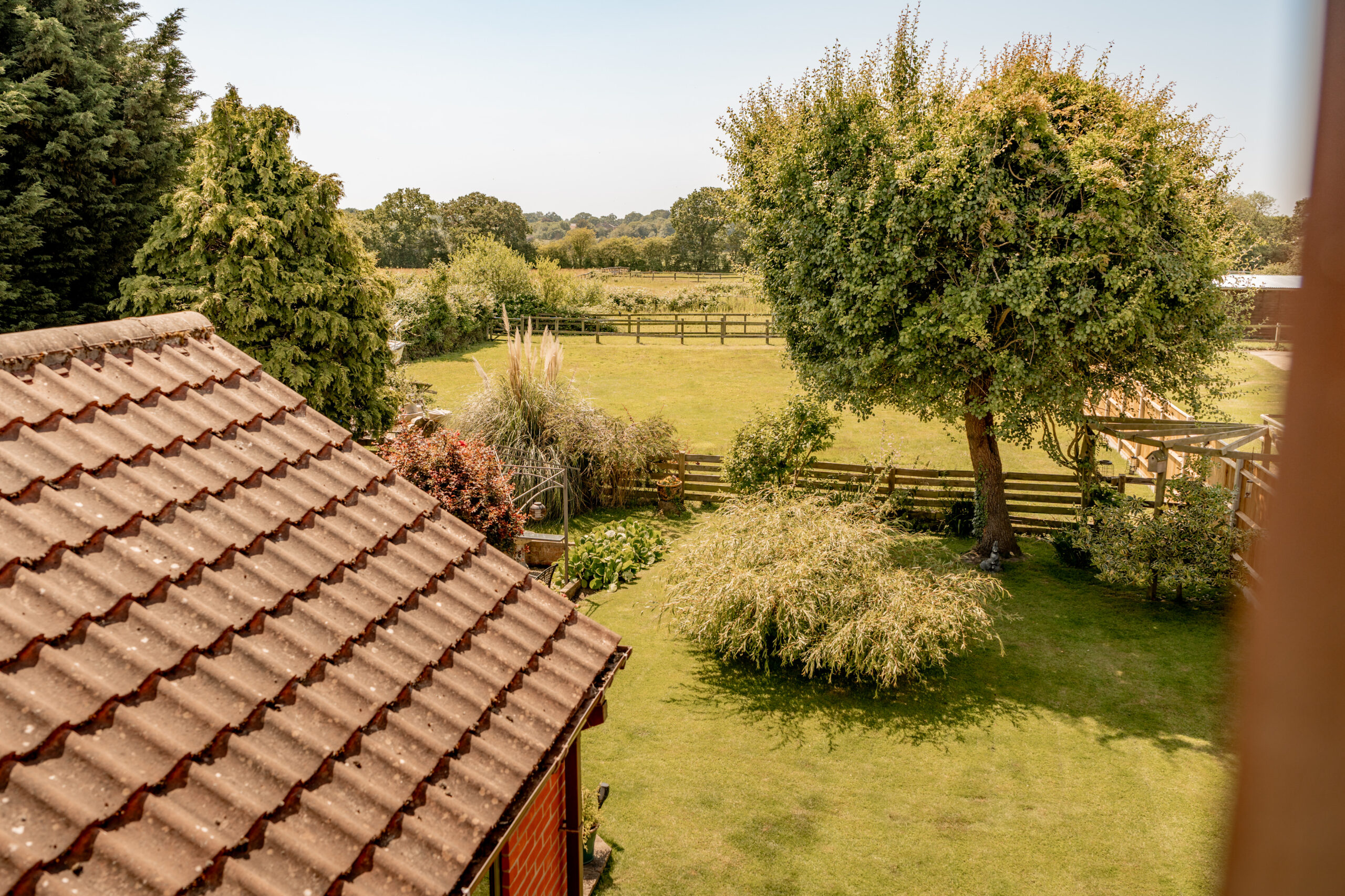 View of a backyard garden with a tiled roof in the foreground, a large tree, green lawn, fence, and open fields with trees and bushes in the distance under a clear sky.