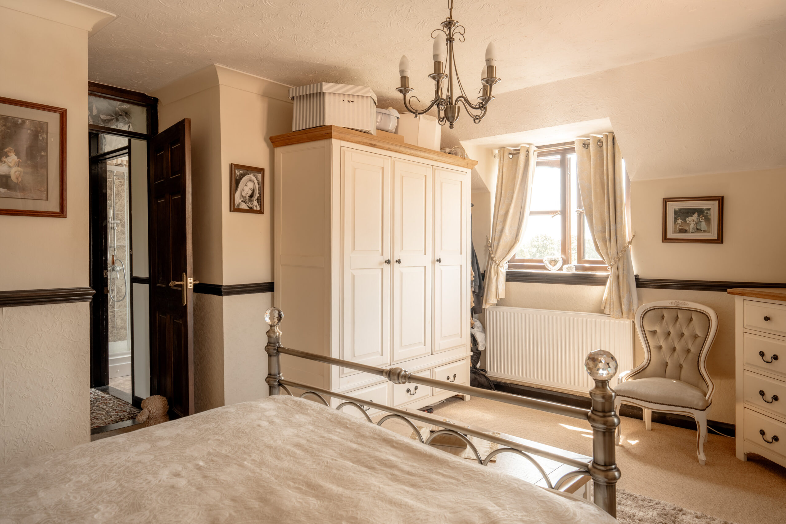 A cozy, sunlit bedroom with a metal bed frame, white wardrobe, tufted chair, dresser, chandelier, two framed pictures, and a window with cream curtains letting in natural light.
