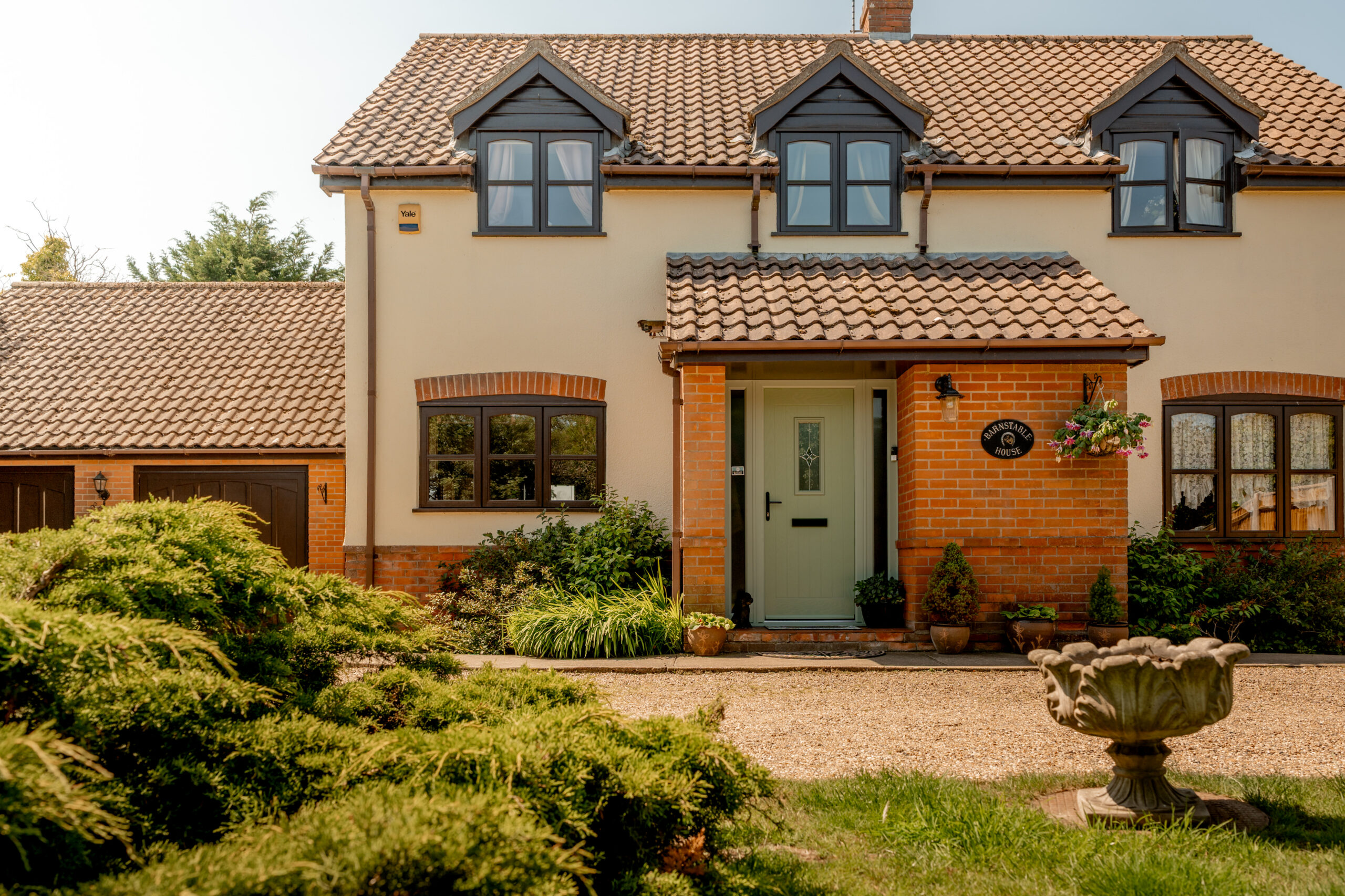 A two-story house with a tiled roof, cream exterior, and brick accents. There are dark-framed windows, a green front door, and a hanging flower basket. A gravel path and a birdbath are in the landscaped front yard.