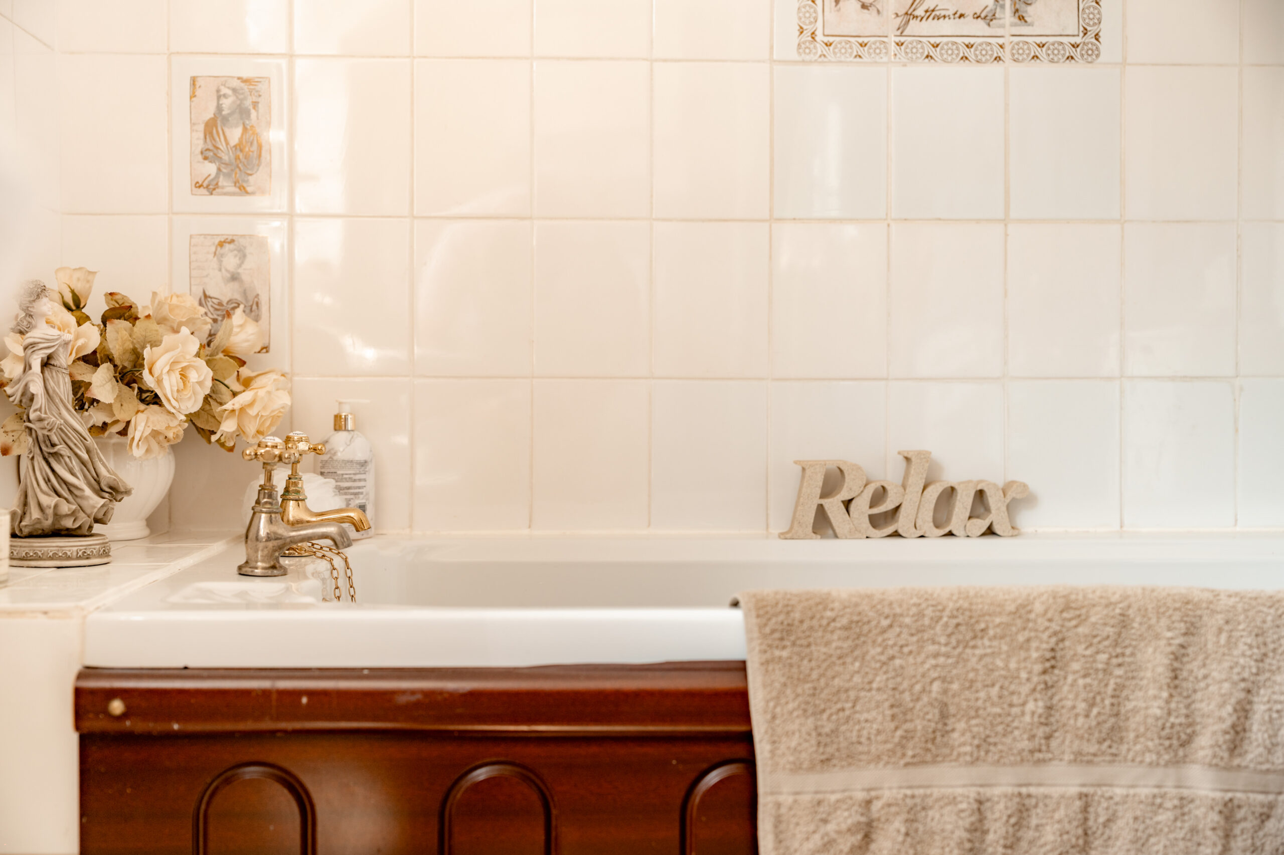 A bathtub with a “Relax” sign on the edge, a silver faucet, a beige towel draped over the side, decorative flowers, and lotion on a tiled wall background.